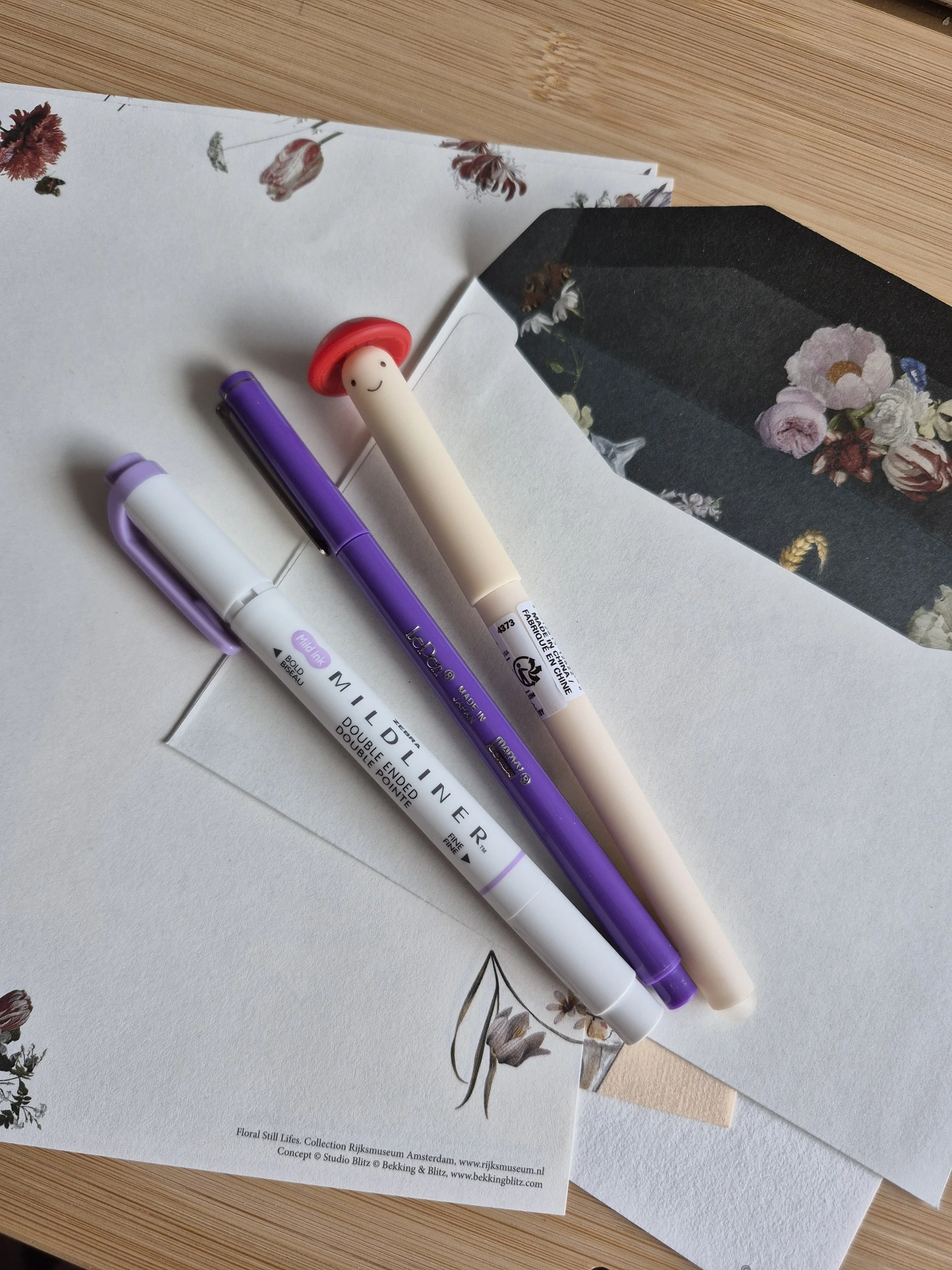 Various stationery items including white, purple, and cream-colored pens resting on decorative paper with floral and botanical prints on a wooden surface.