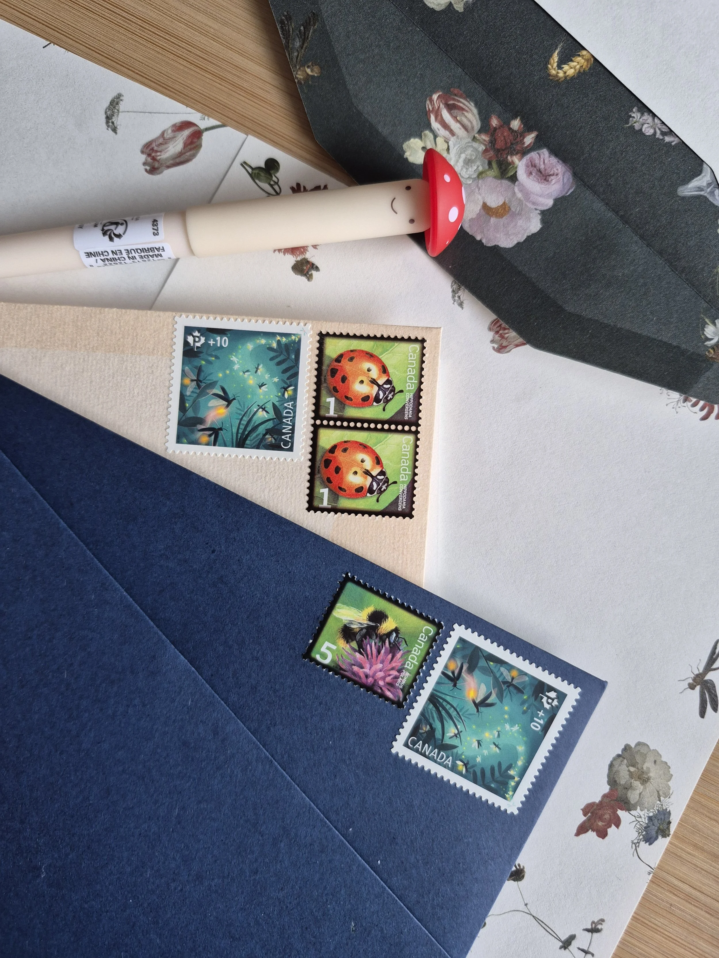 A flat lay of Canadian postage stamps featuring nature and insects, with a white and red mushroom-shaped pen, on top of floral-patterned paper and blue and beige envelopes.
