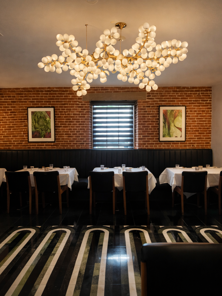 A modern dining room with a brick wall, a window with blinds, and a chandelier with white globes. Eight tables are set with white tablecloths and glasses, surrounded by black chairs. The floor has a black, white, and green patterned tile.