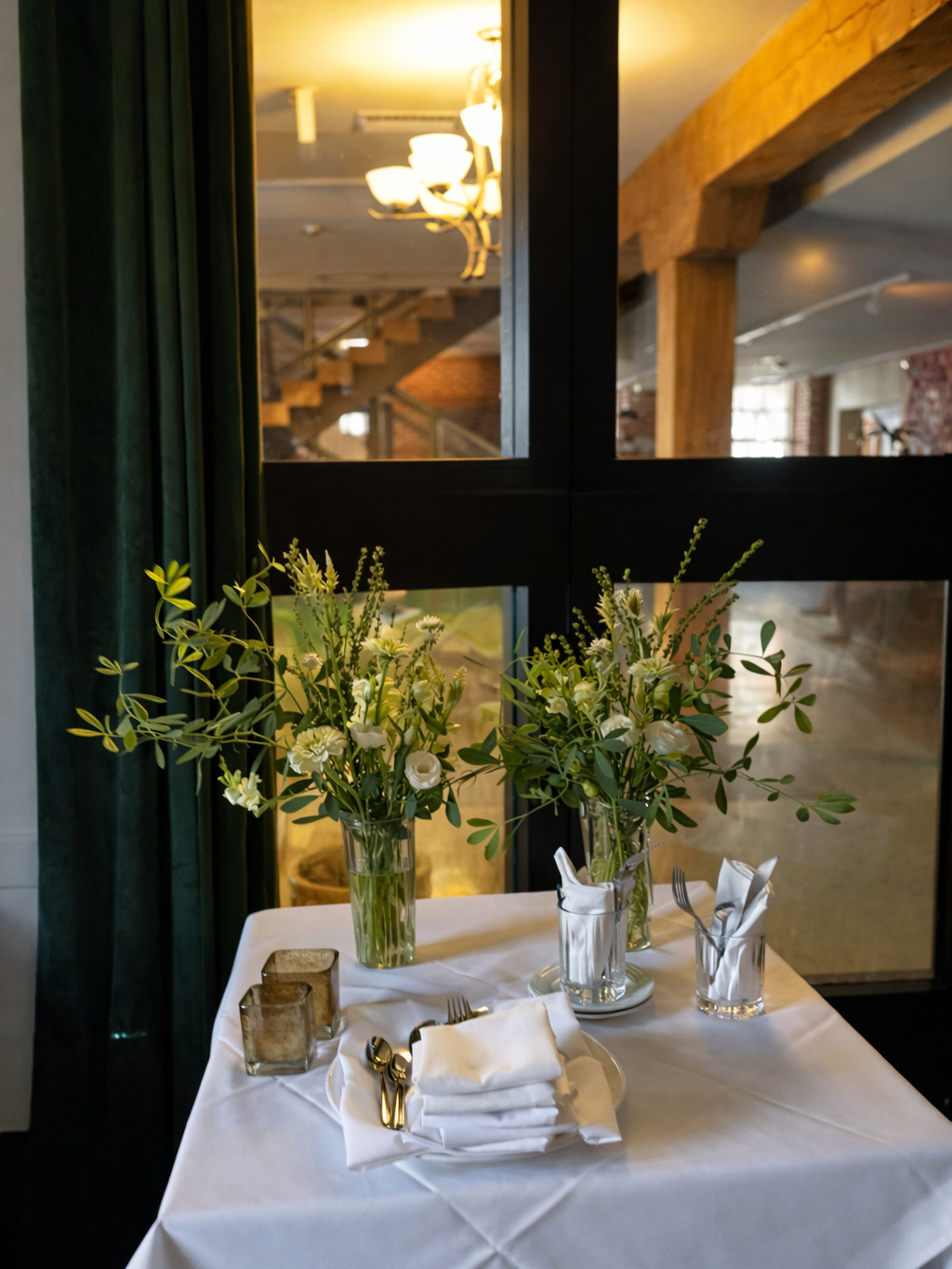 A table set for dining with white napkins, silverware, and glassware, decorated with three vases of white flowers and green foliage in a restaurant with large windows and wooden beams.