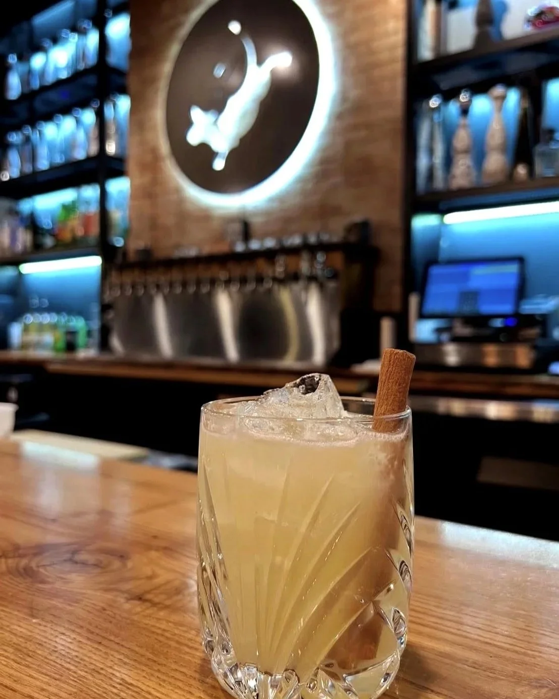 A cocktail with ice and a cinnamon stick in a glass on a wooden bar counter, with a blurred background of a bar with shelves, a large clock with a cat on the wall, and a cash register.