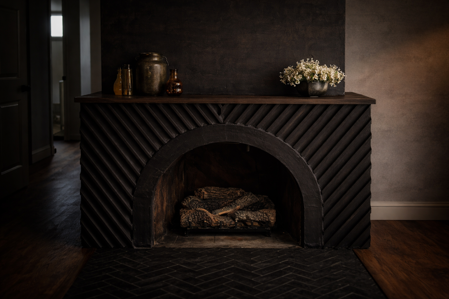 A dark fireplace with a wooden mantel decorated with vases and a black bowl of white flowers, on a dark wall. The fireplace has a black wood frame with diagonal slats, a rounded opening, and logs inside, resting on a black brick hearth.