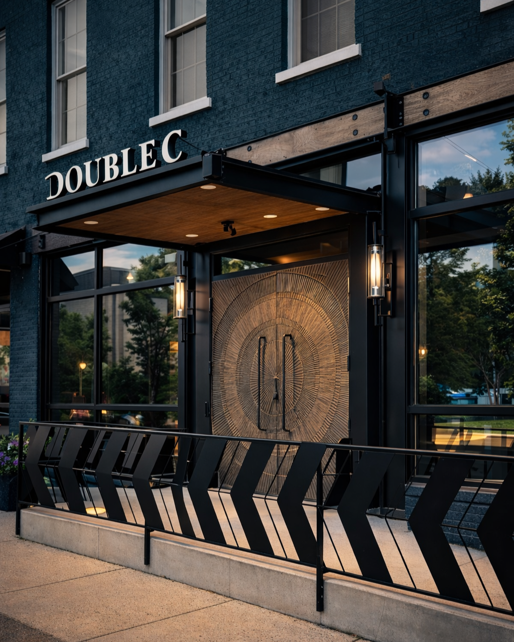 Modern building with a sign reading 'Double C' above the entrance, featuring large glass windows, black metal framing, and decorative lighting, with trees reflected in the window.