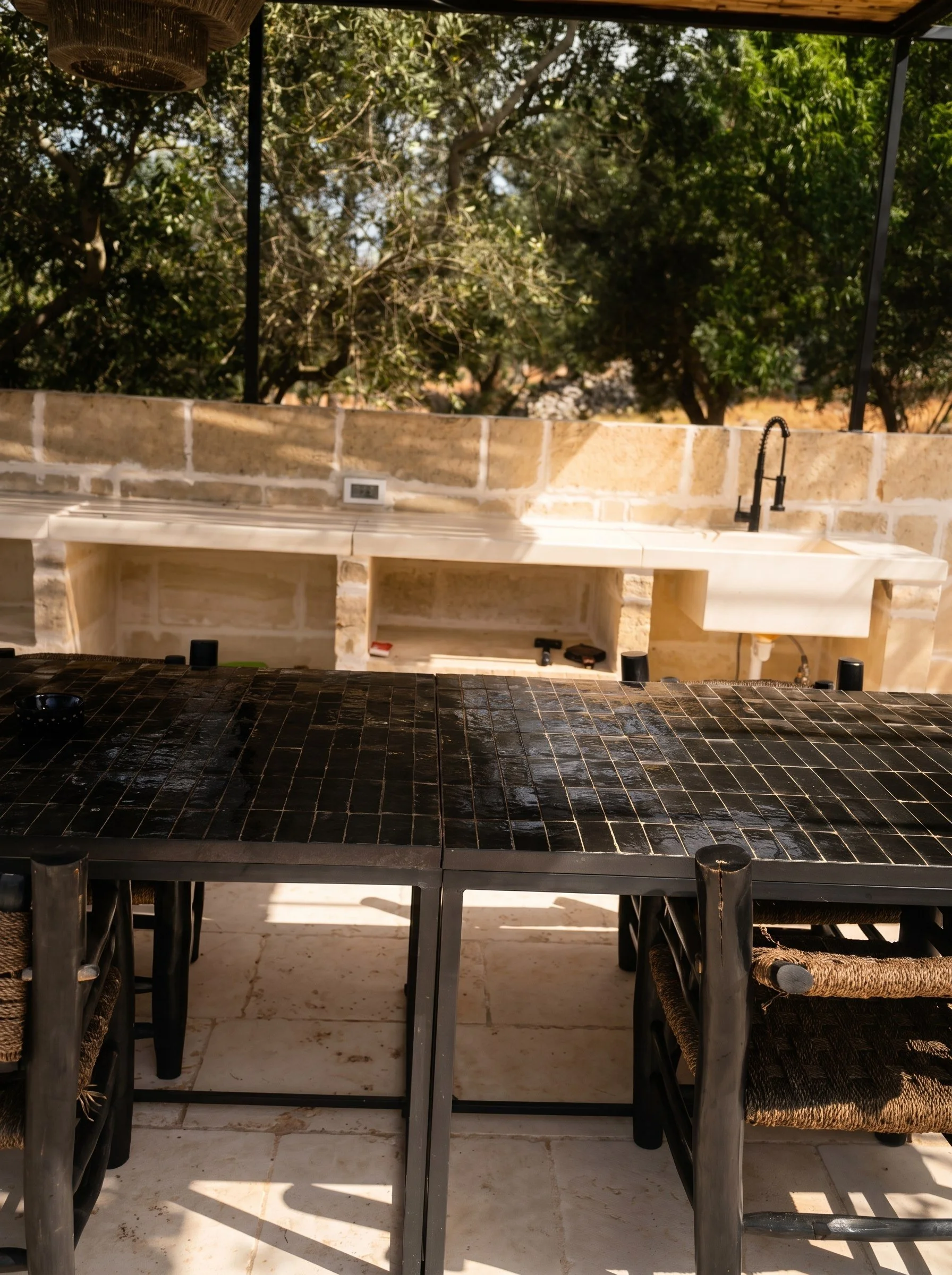 Outdoor kitchen with a black tiled counter, a brick wall with a sink and black faucet, and a view of trees in the background.