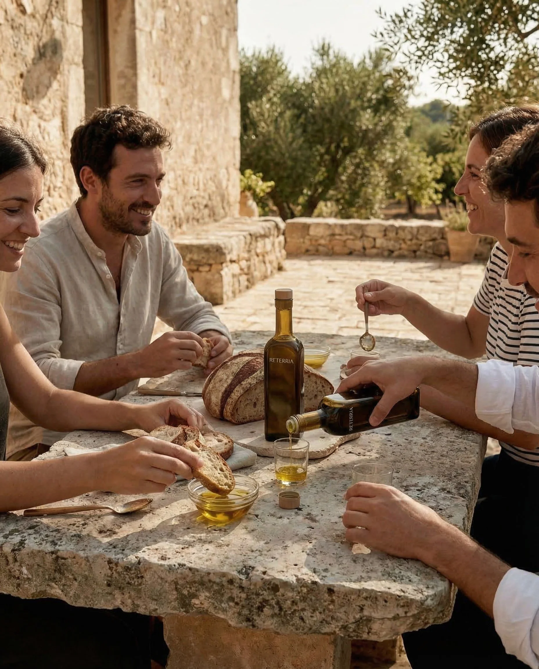 Group of people sitting around a stone table outdoors, eating bread and enjoying olive oil, with a stone building and trees in the background on a sunny day.