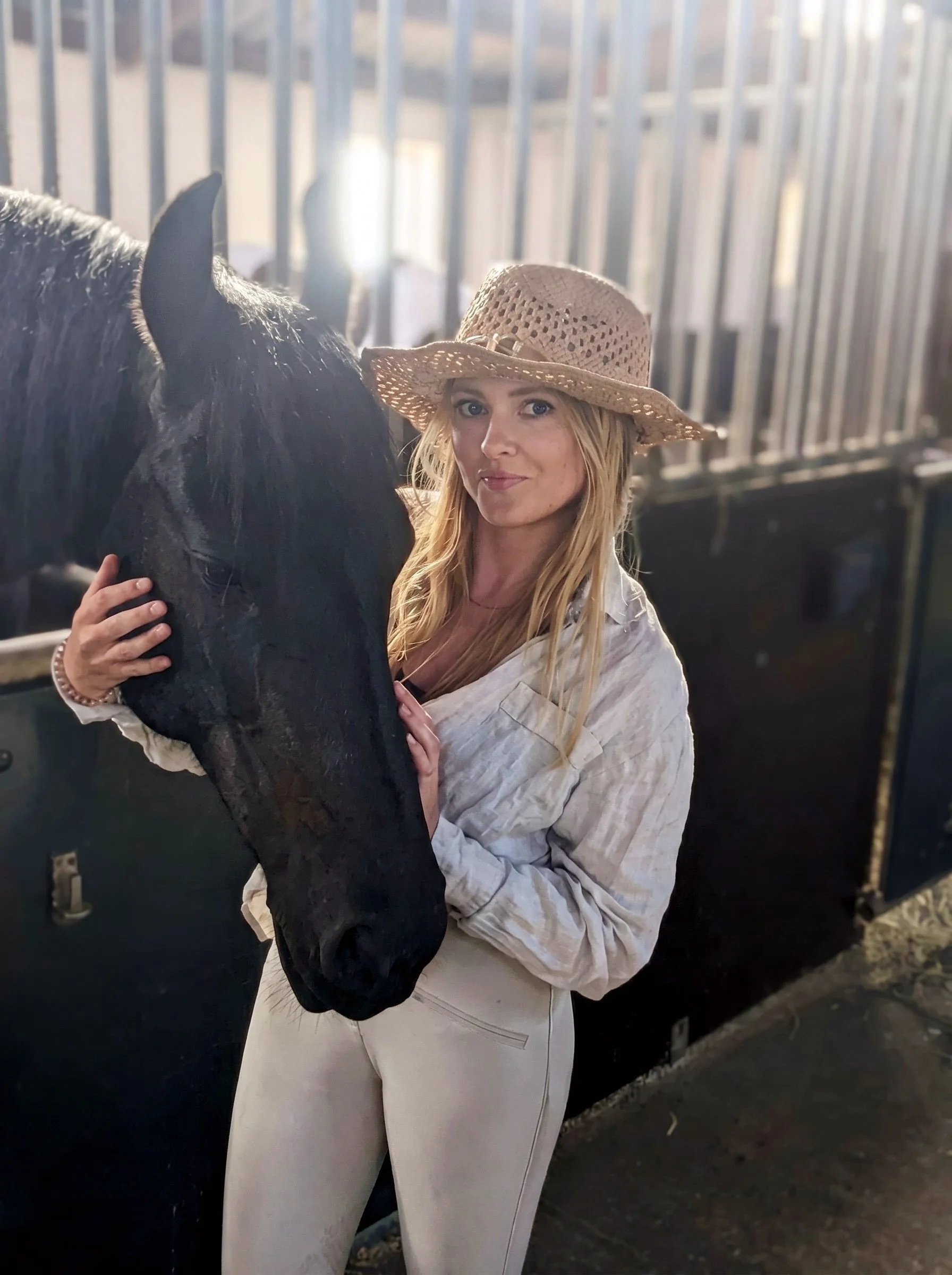 A woman with long blonde hair wearing a straw hat and a light-colored jacket standing near a black horse behind a metal fence.