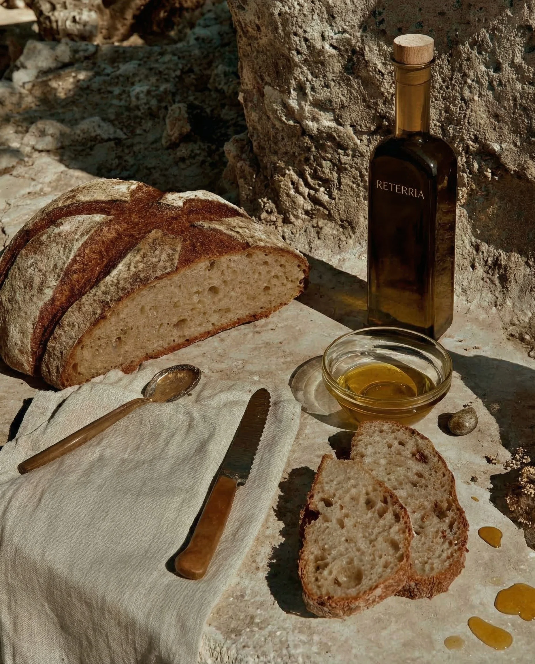 Sliced bread on a cloth, a knife, a jar of olive oil, and a glass bowl of olive oil on a rustic surface with a stone background.