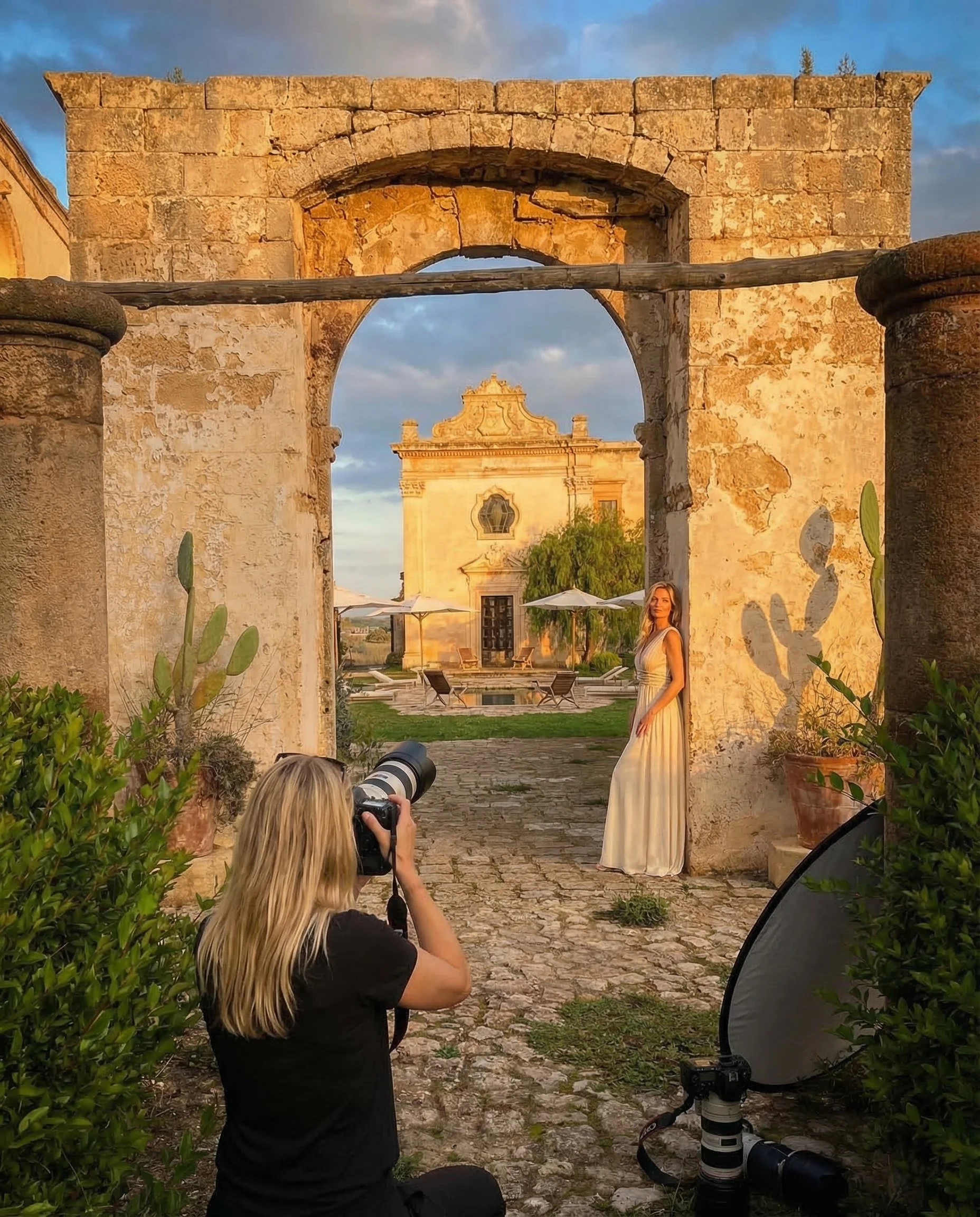 A woman in a white dress posing in front of an ancient stone archway during sunset with a photographer taking her picture. The setting includes greenery, stone pavement, and historic architecture in the background.