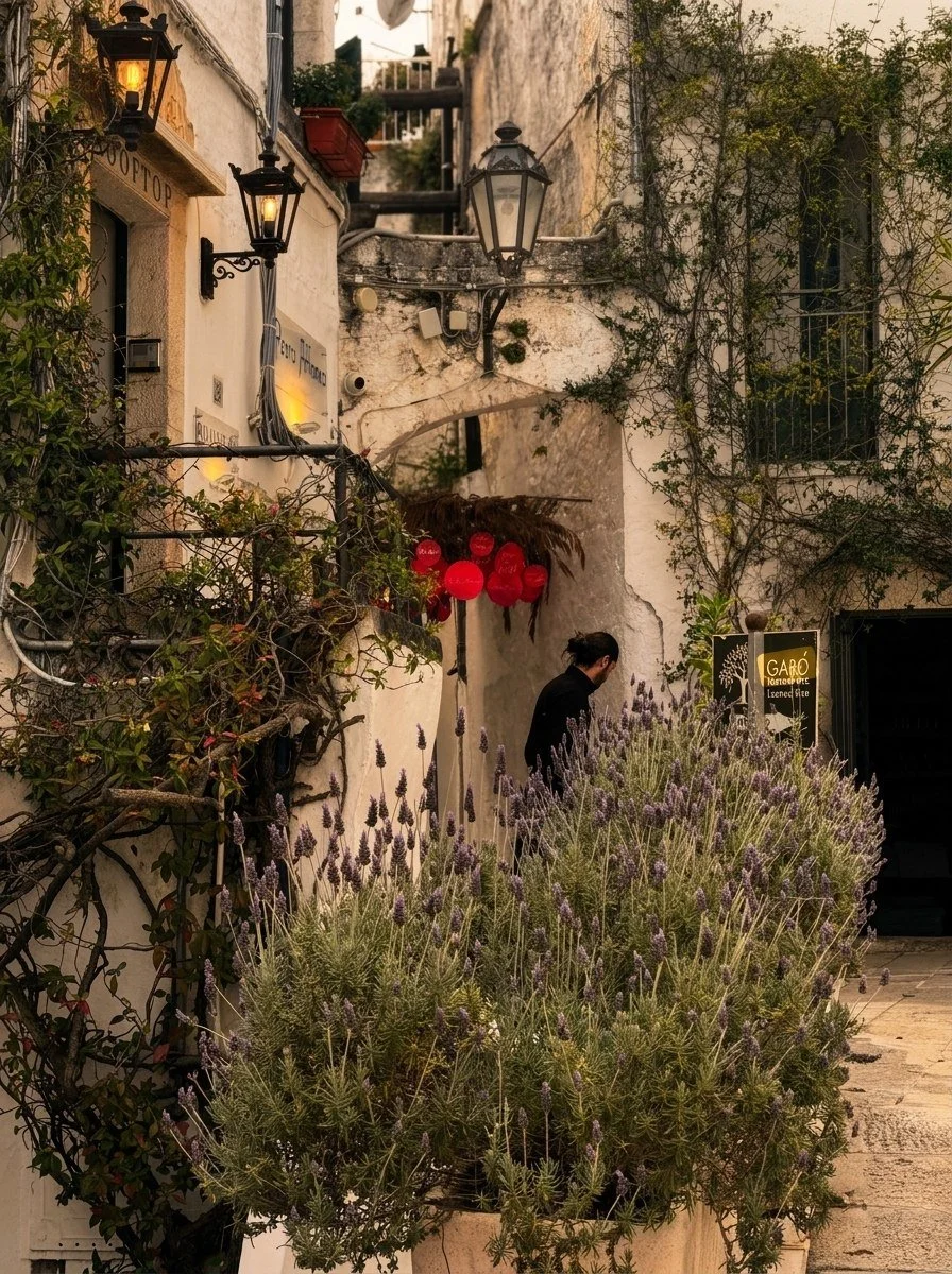A narrow alleyway with white stucco buildings, decorative outdoor lanterns, and climbing plants. There are purple flowering plants in the foreground, and a person with dark hair in a bun standing near a sign for a restaurant called Garó.
