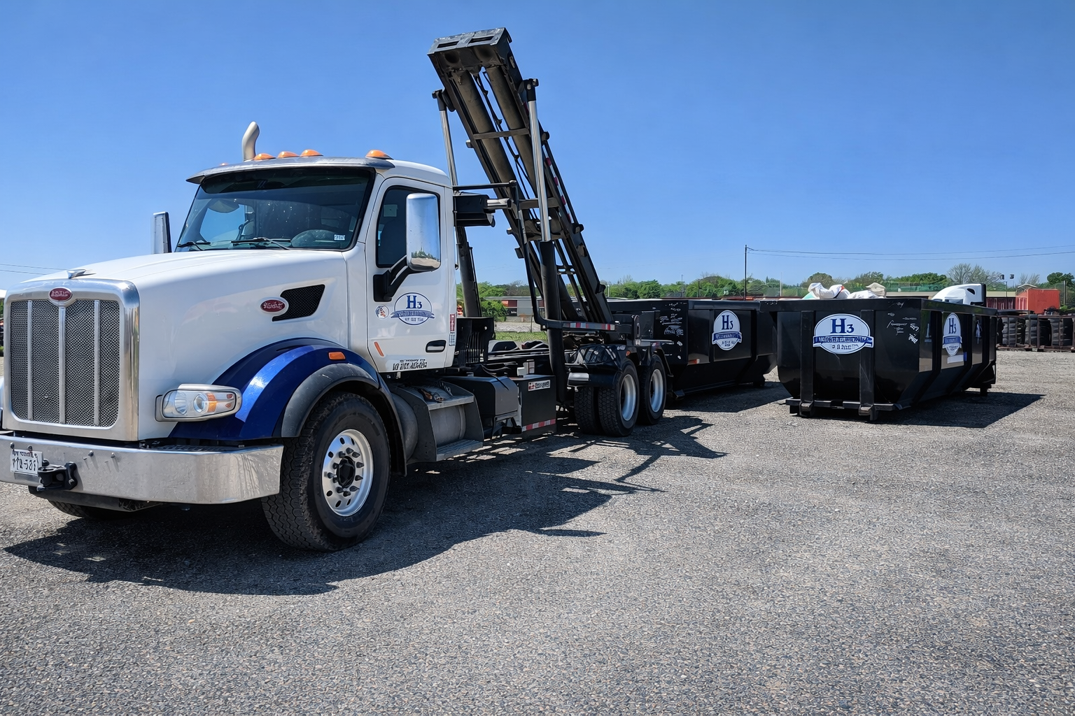 White and blue flatbed truck with a crane arm, parked on gravel, with large black containers and a clear blue sky in the background.