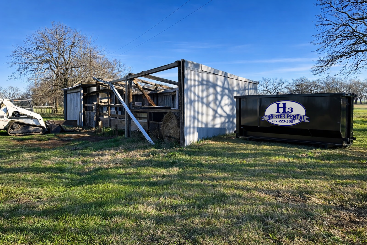 A damaged shed with leaning and broken wooden parts, an abandoned backhoe, and a black dumpster labeled 'H3 Dumpster Rental' on a grassy field with trees and blue sky in the background.