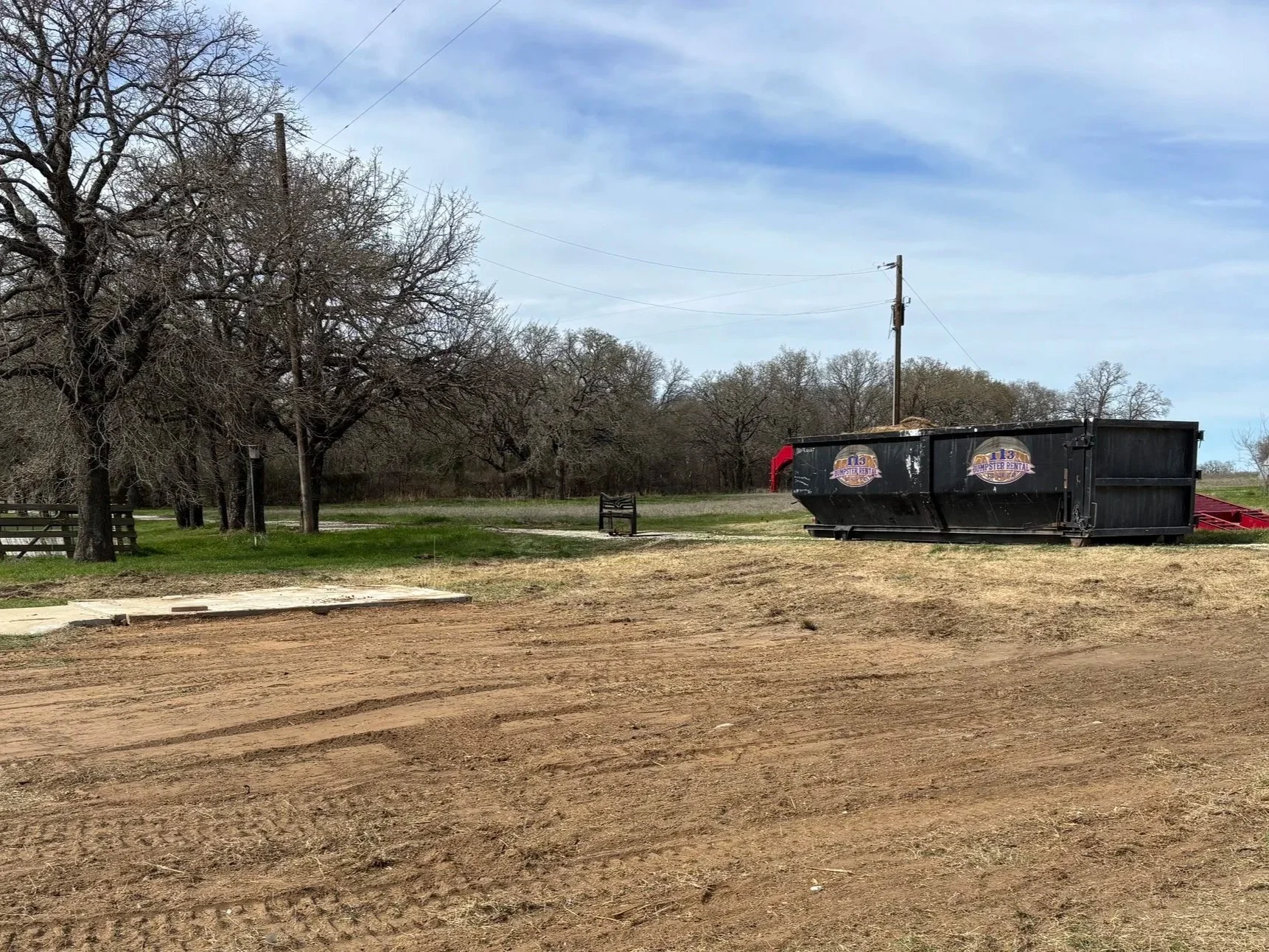 A rural scene with a dirt road, leafless trees, two park benches, and a large black dumpster with a sign on it, under a partly cloudy sky.