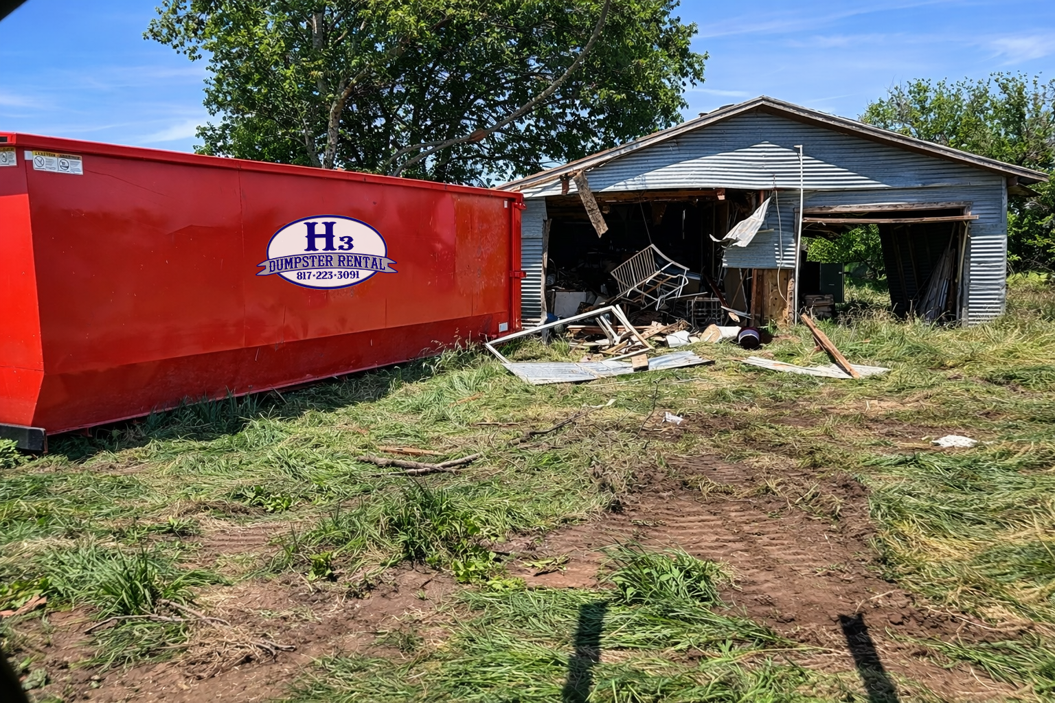 A damaged house with a partially collapsed front wall, debris and broken furniture inside, and a large red dumpster with the label H3 Dumpster Rental parked in front. The scene shows signs of a recent storm or disaster, with a clear blue sky and green trees in the background.