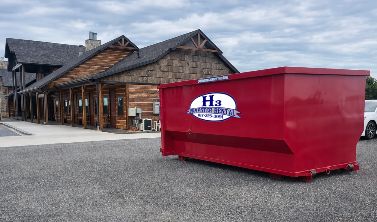 A large red dumpster with a sign for H3 Dumpster Rental parked in front of a wooden building with a covered porch.