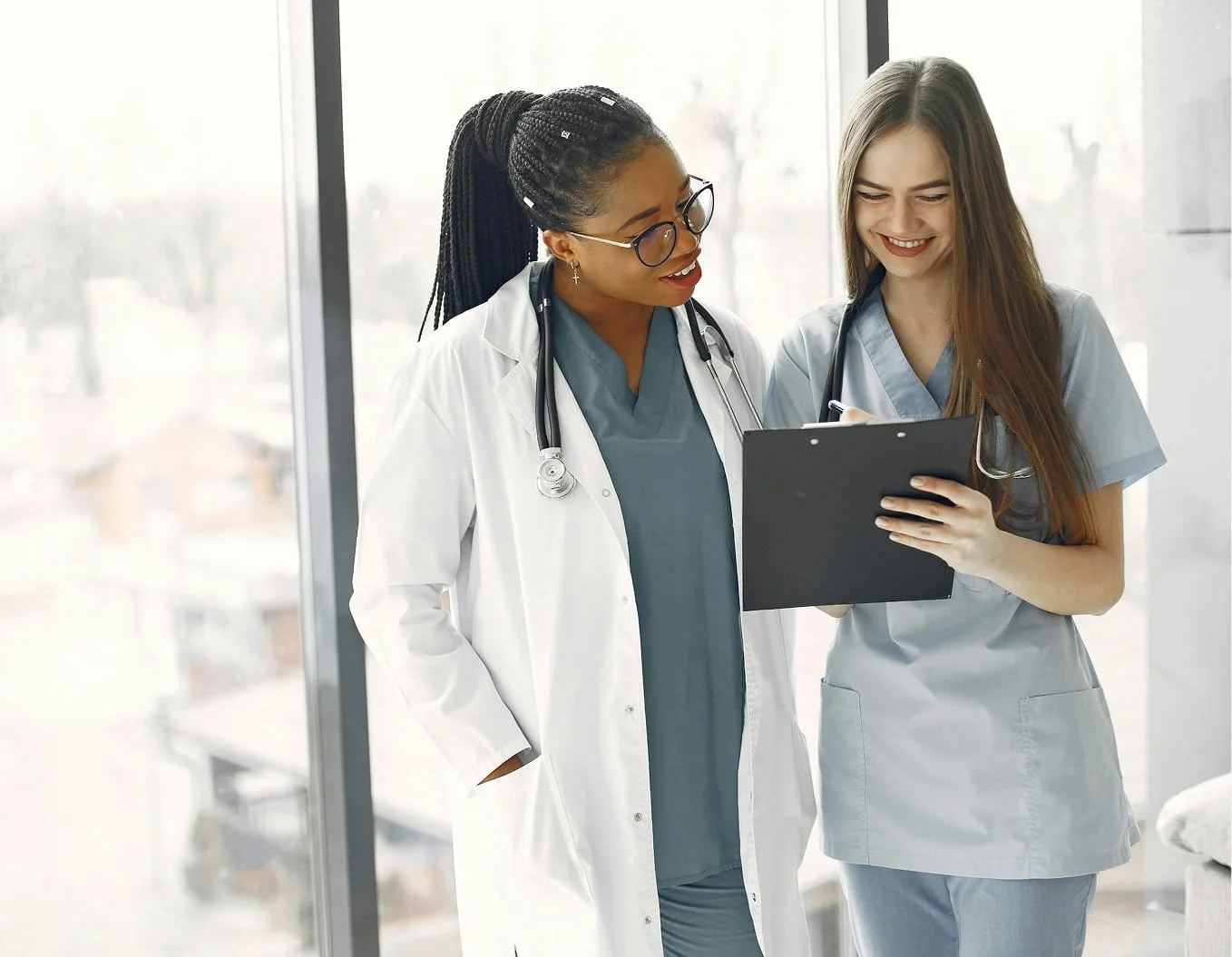 Two female healthcare professionals, one in a white doctor's coat and the other in blue scrubs, reviewing a clipboard together in a bright medical facility.