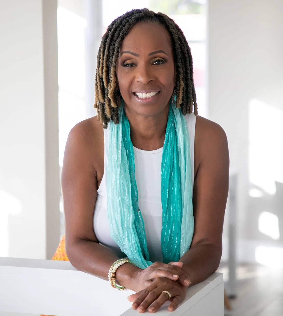 A smiling woman with dark skin and stylish dreadlocks, wearing a white sleeveless top and a turquoise scarf, sitting with her hands clasped on a white table in a bright room with natural light.