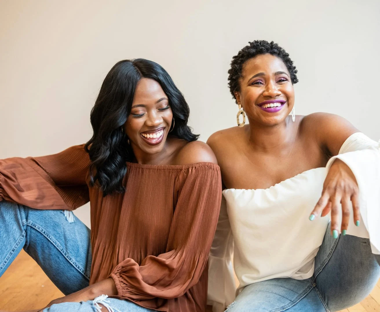 Two women sitting on the floor, smiling and laughing together, one wearing an off-shoulder brown top and the other in a white off-shoulder top.