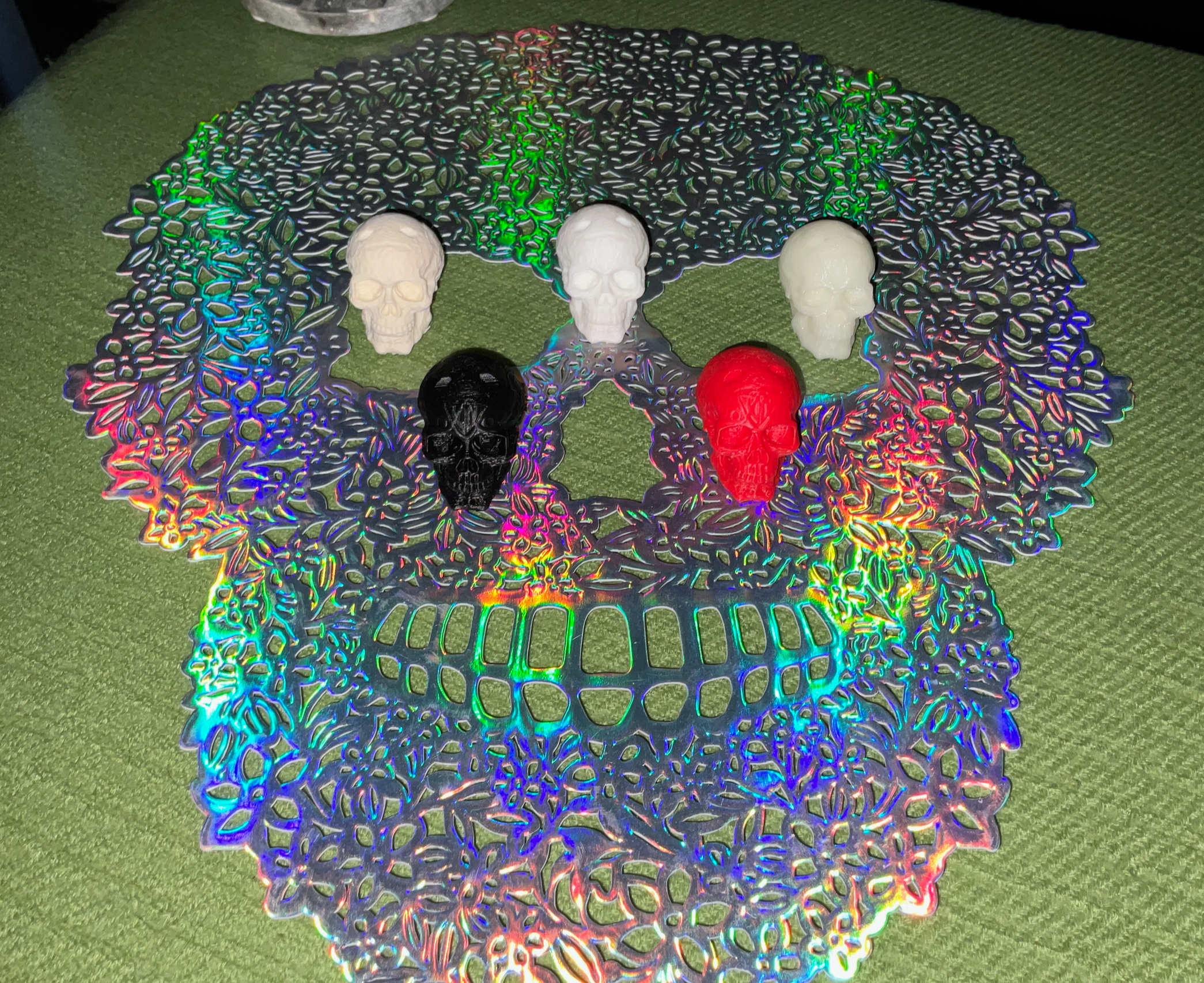 Reflective mat (not printed but we can!) showcasing 3D-printed skulls in PLA, PETG, ASA, ABS, and TPU on a green surface. Showcasing our various filaments.