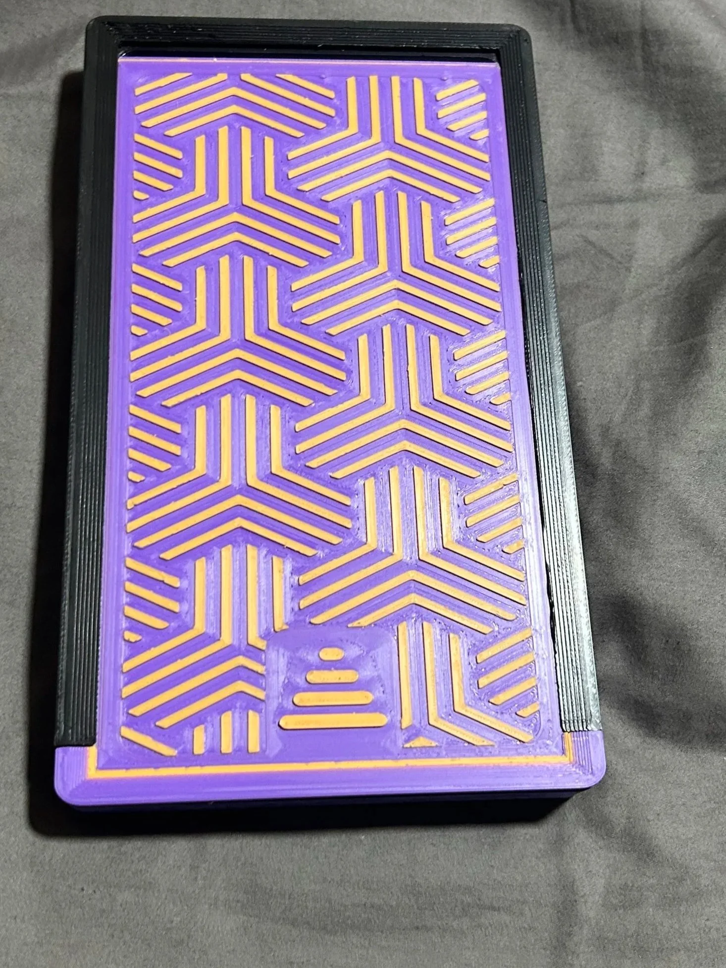 A purple rectangular object with an intricate yellow geometric pattern consisting of connected chevrons and lines, bordered by a black frame.