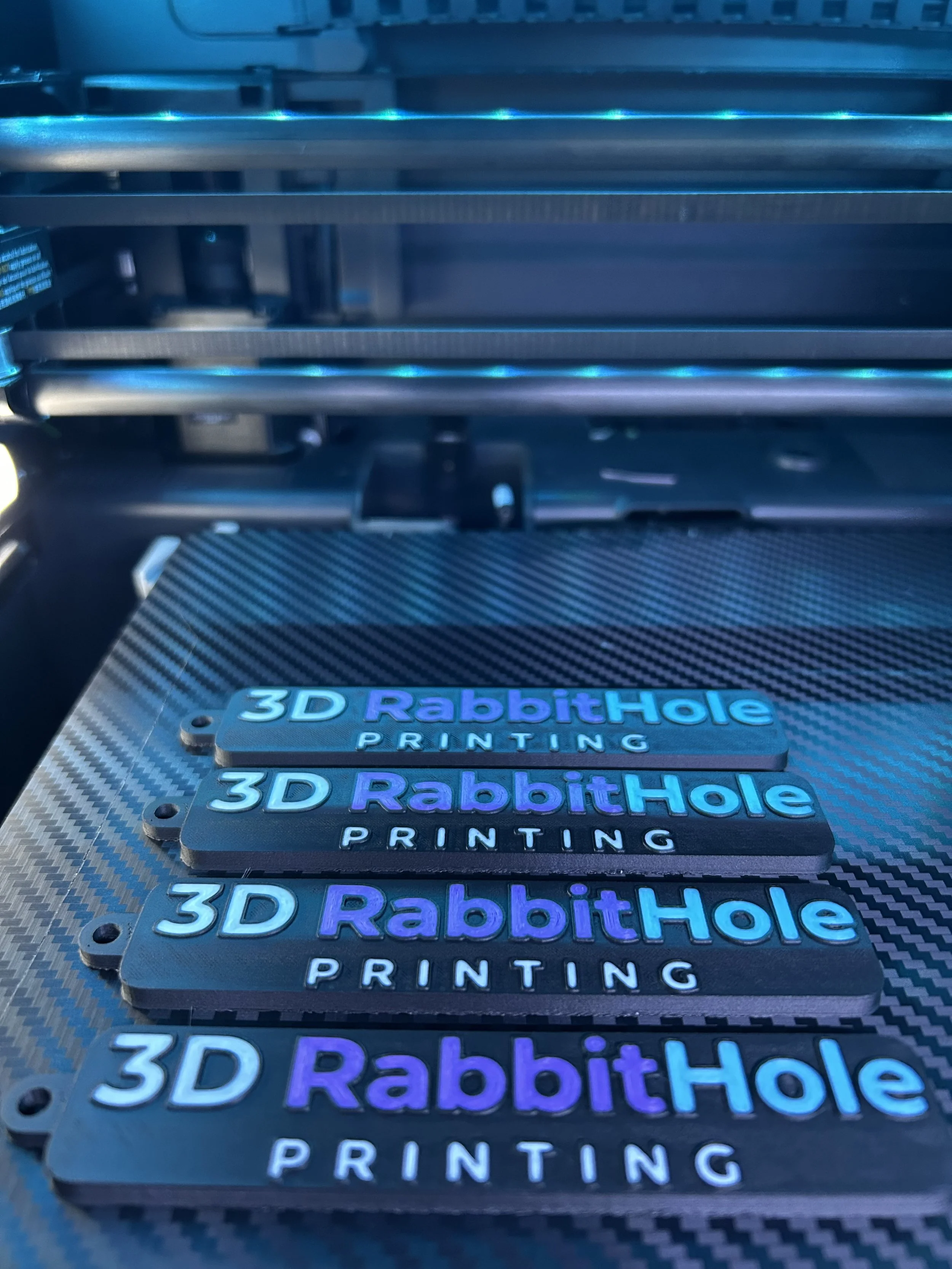 Four black keychains with the text '3D Rabbit Hole PRINTING' in blue, purple, and white lettering, placed on a textured surface, with a 3D printer in the background.