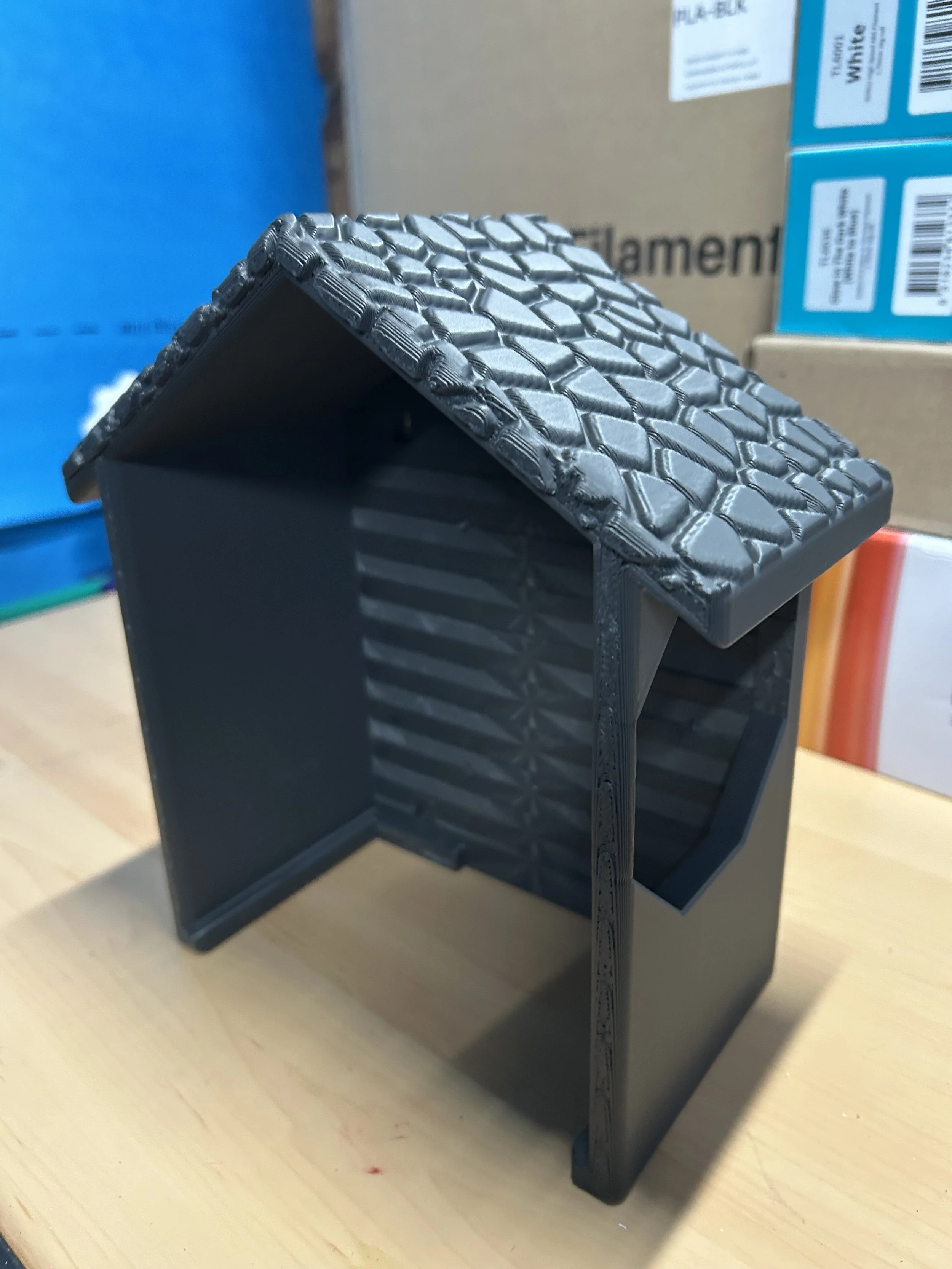 A black plastic miniature house with a textured roof, situated on a wooden surface.