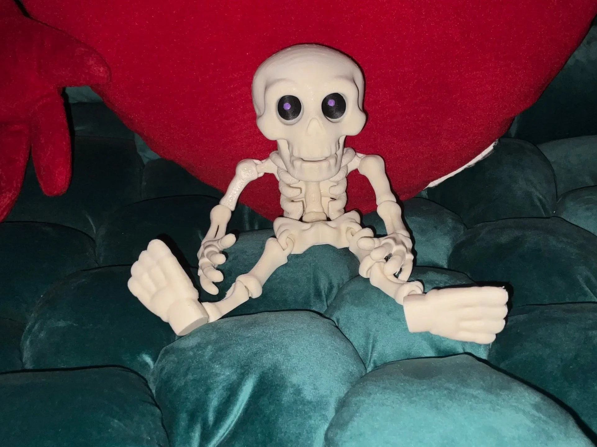 A small plastic skeleton toy with large black and purple eyes, sitting on a green tufted surface, in front of a red cushion and a red plush object.