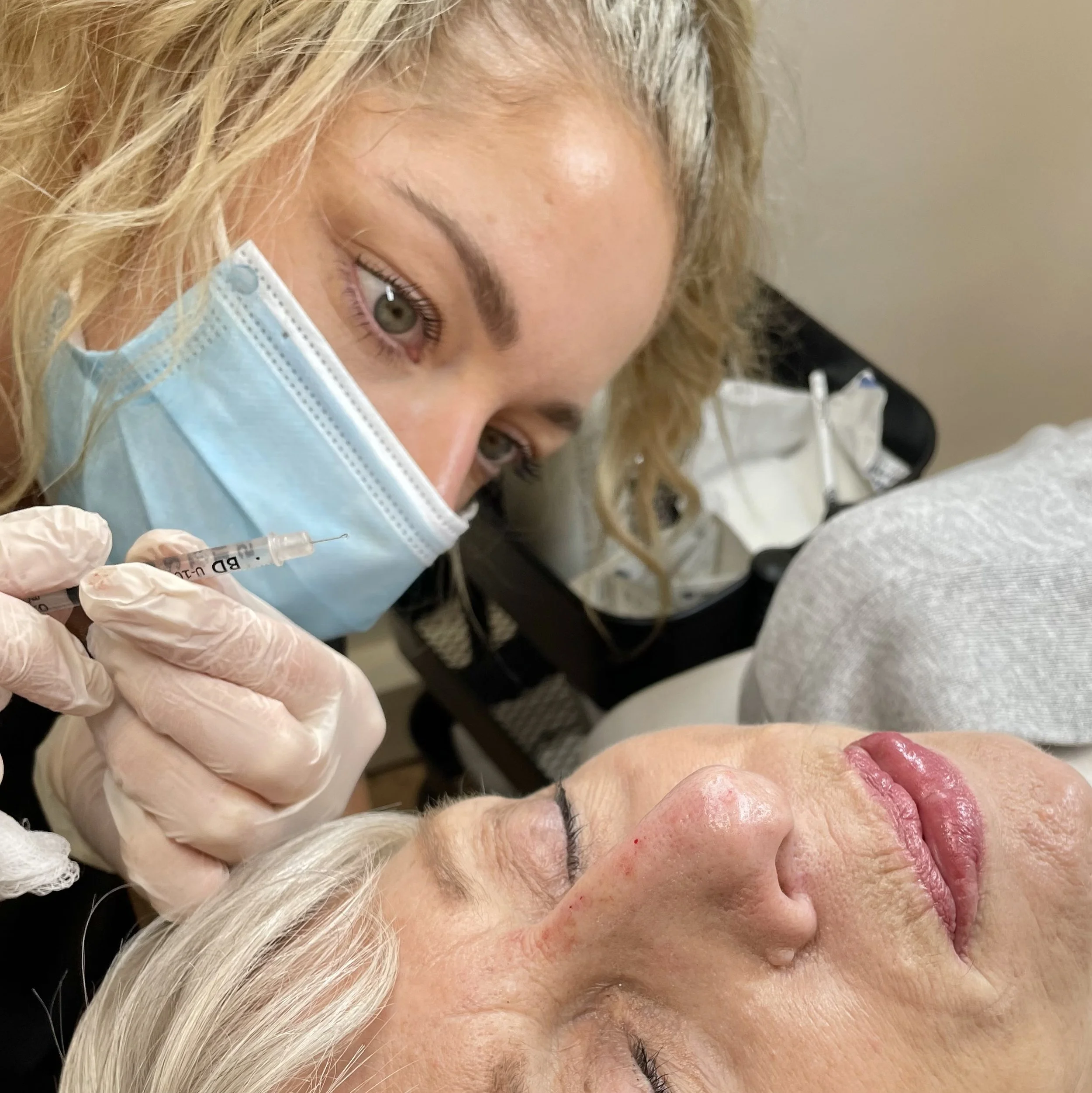 A healthcare professional in a surgical mask and gloves administers a cosmetic injection to an elderly woman's face, who is lying down with eyes closed.