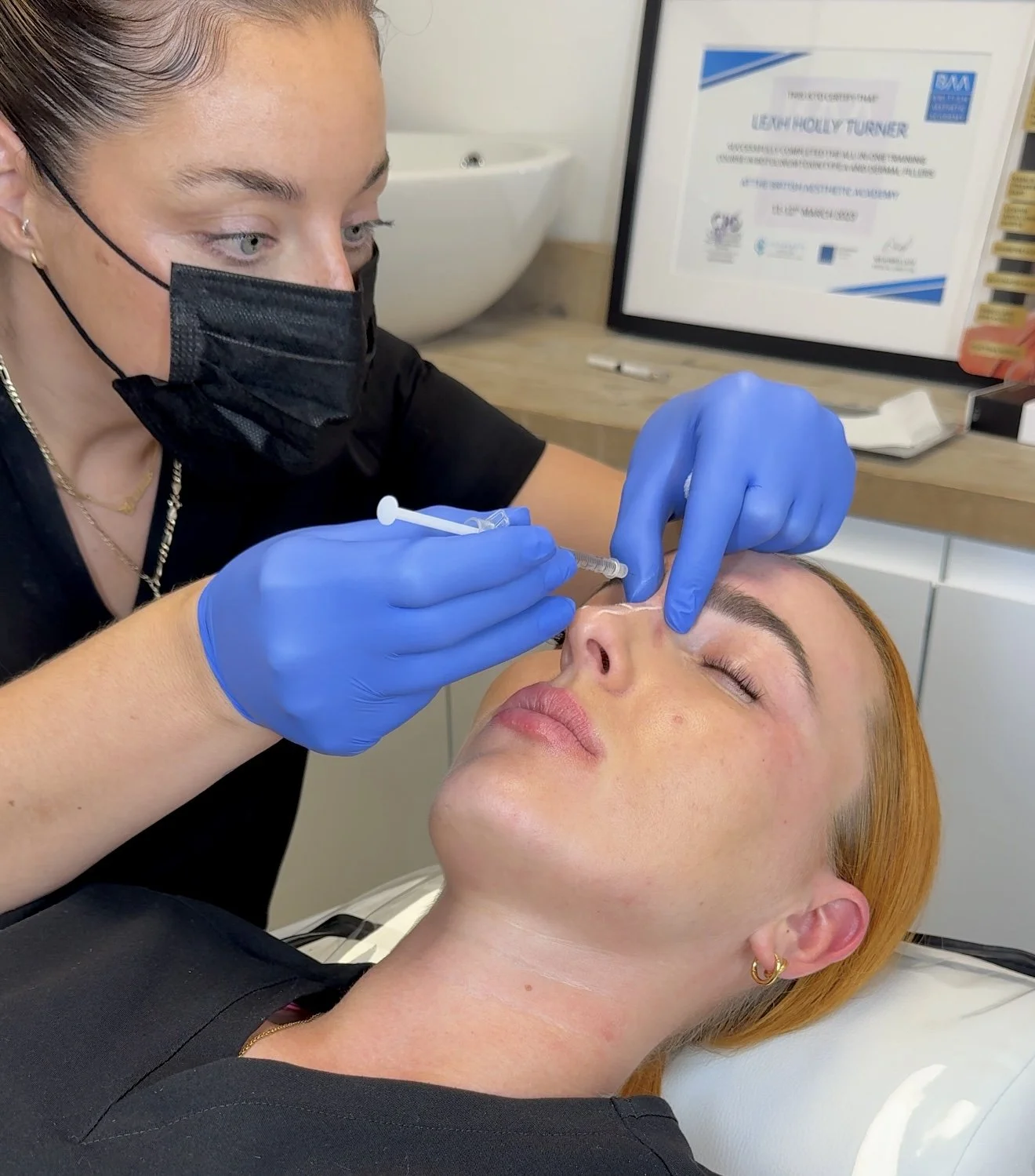 A woman is receiving a cosmetic injection in her nose from a practitioner wearing blue gloves, a black mask, and black scrubs. The patient has closed eyes and orange hair, and is lying on a medical bed indoors.