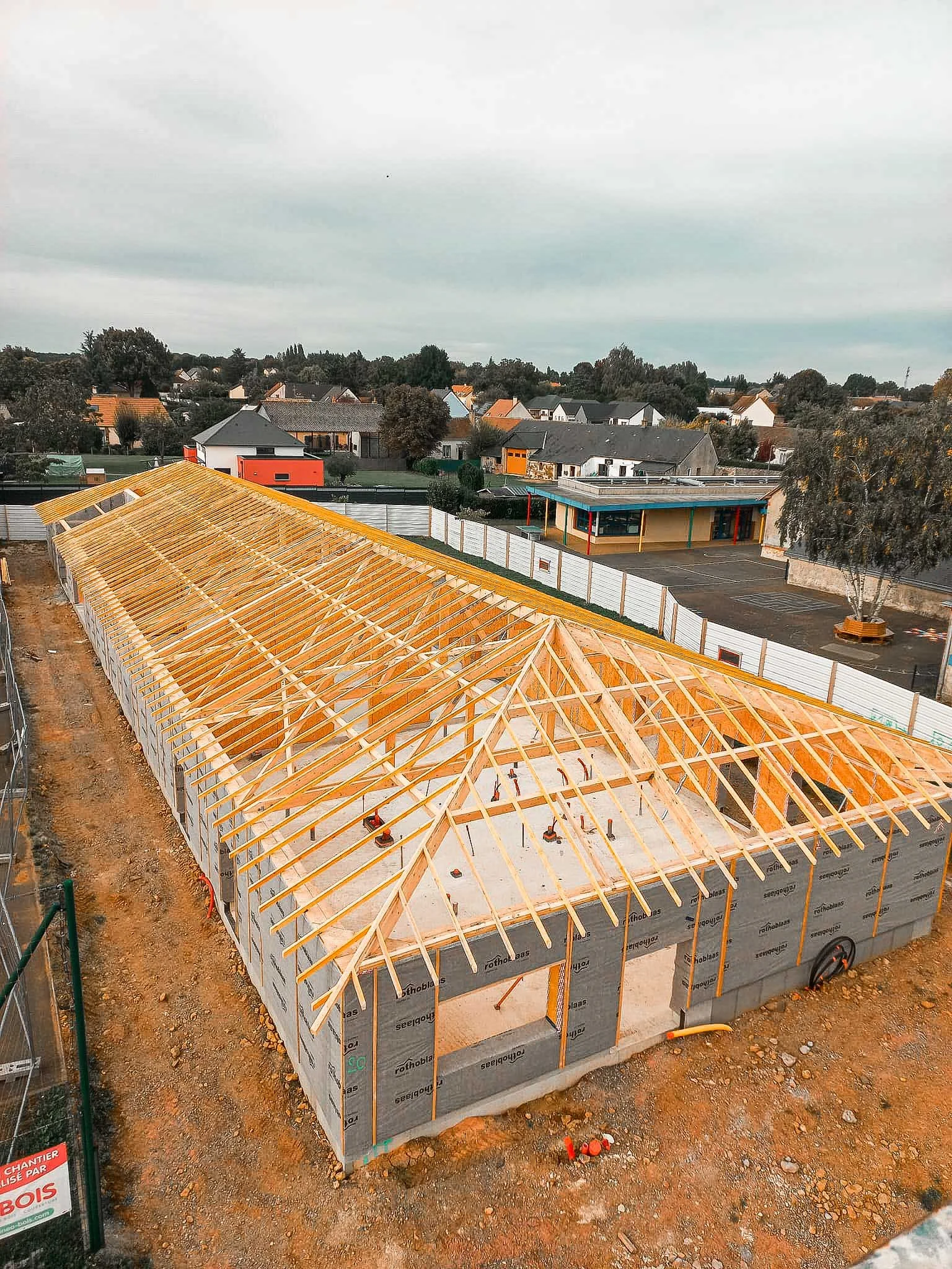 Construction d'un bâtiment avec une structure en bois et un mur en béton, un parking devant et un paysage résidentiel en arrière-plan.