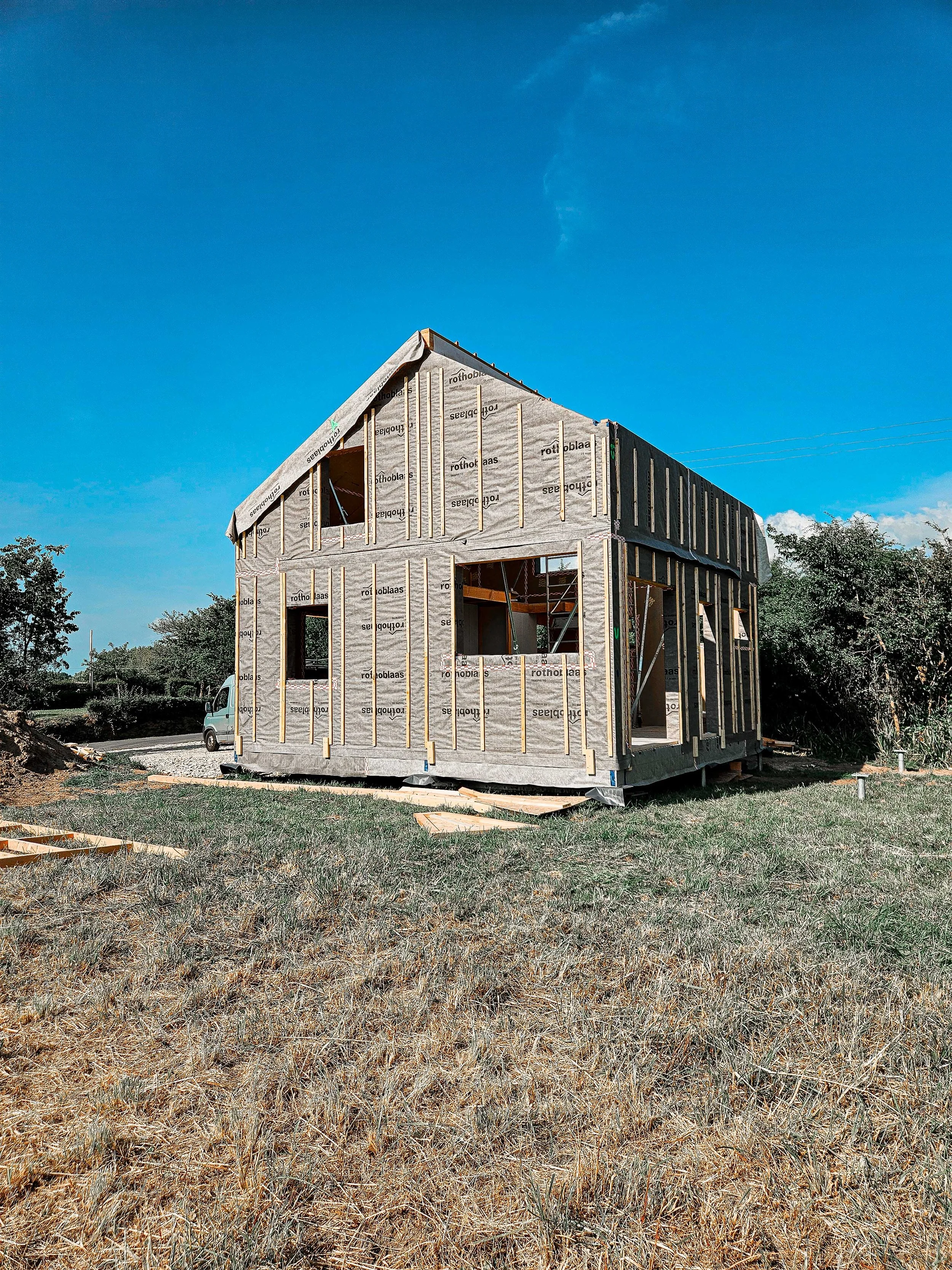 Construction d'une maison en bois avec structure en écorce, sous un ciel bleu clair, entourée de végétation et en cours de construction.