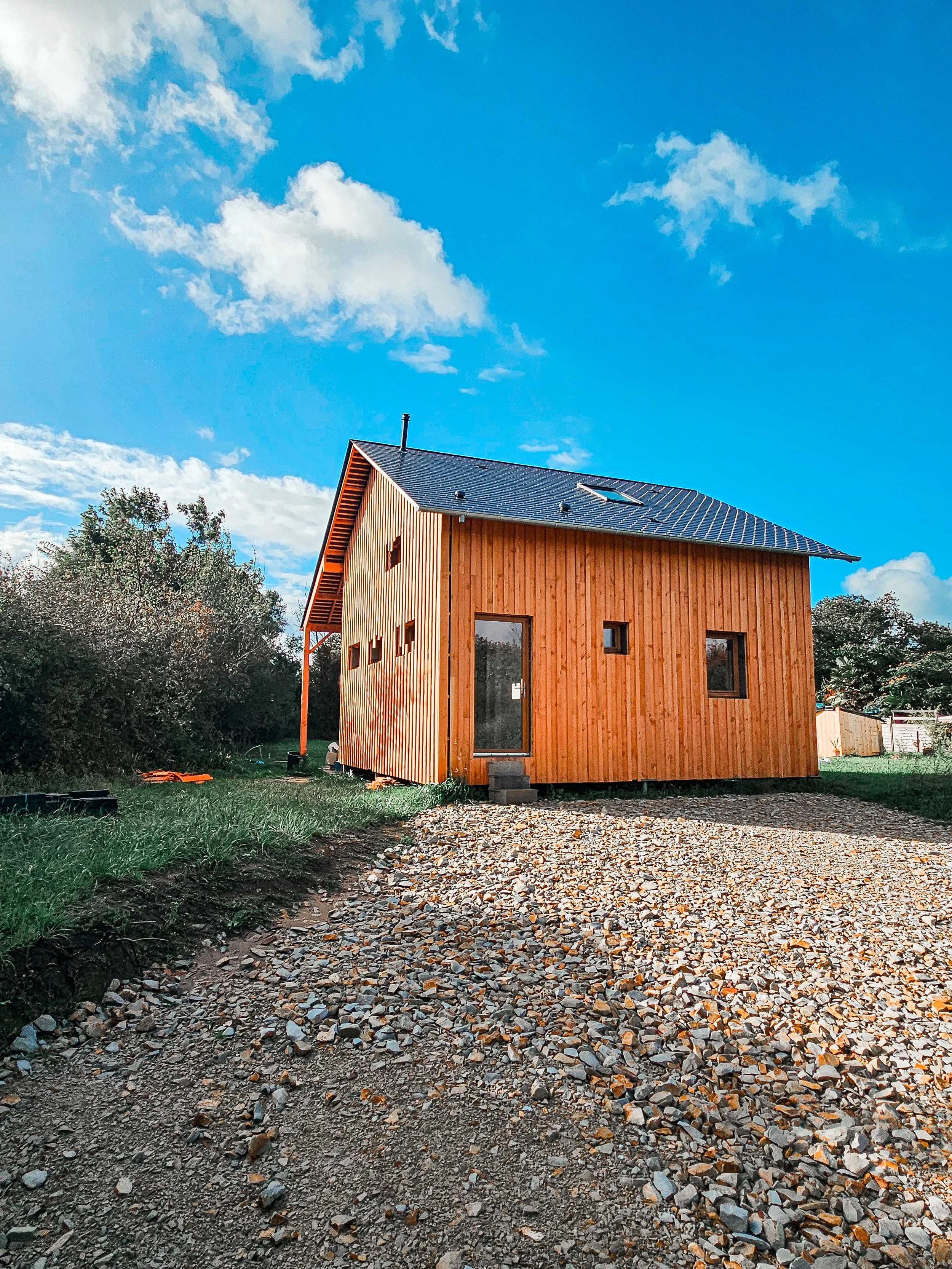 Une maison en bois en construction avec un toit en tuiles bleues, entourée de pelouse et de végétation, sous un ciel bleu avec des nuages.