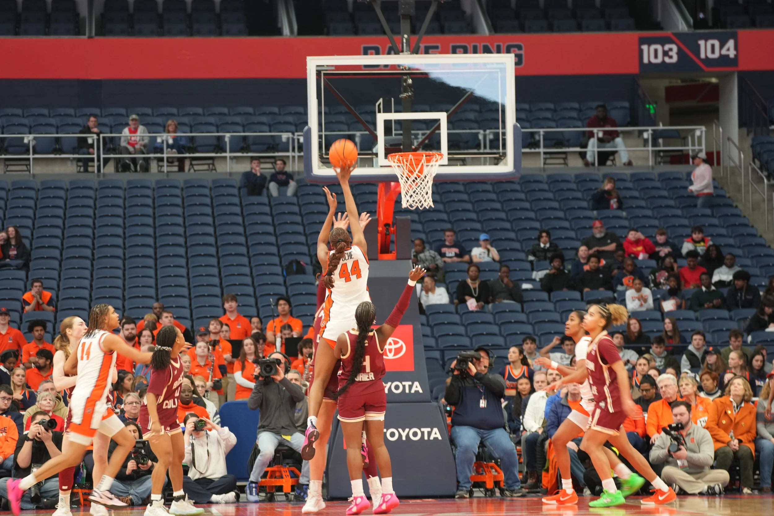 A women's basketball game with a player in a white and orange uniform jumping to shoot the ball towards the basket while a player in a maroon uniform attempts to block. Other players and spectators are visible in the background.