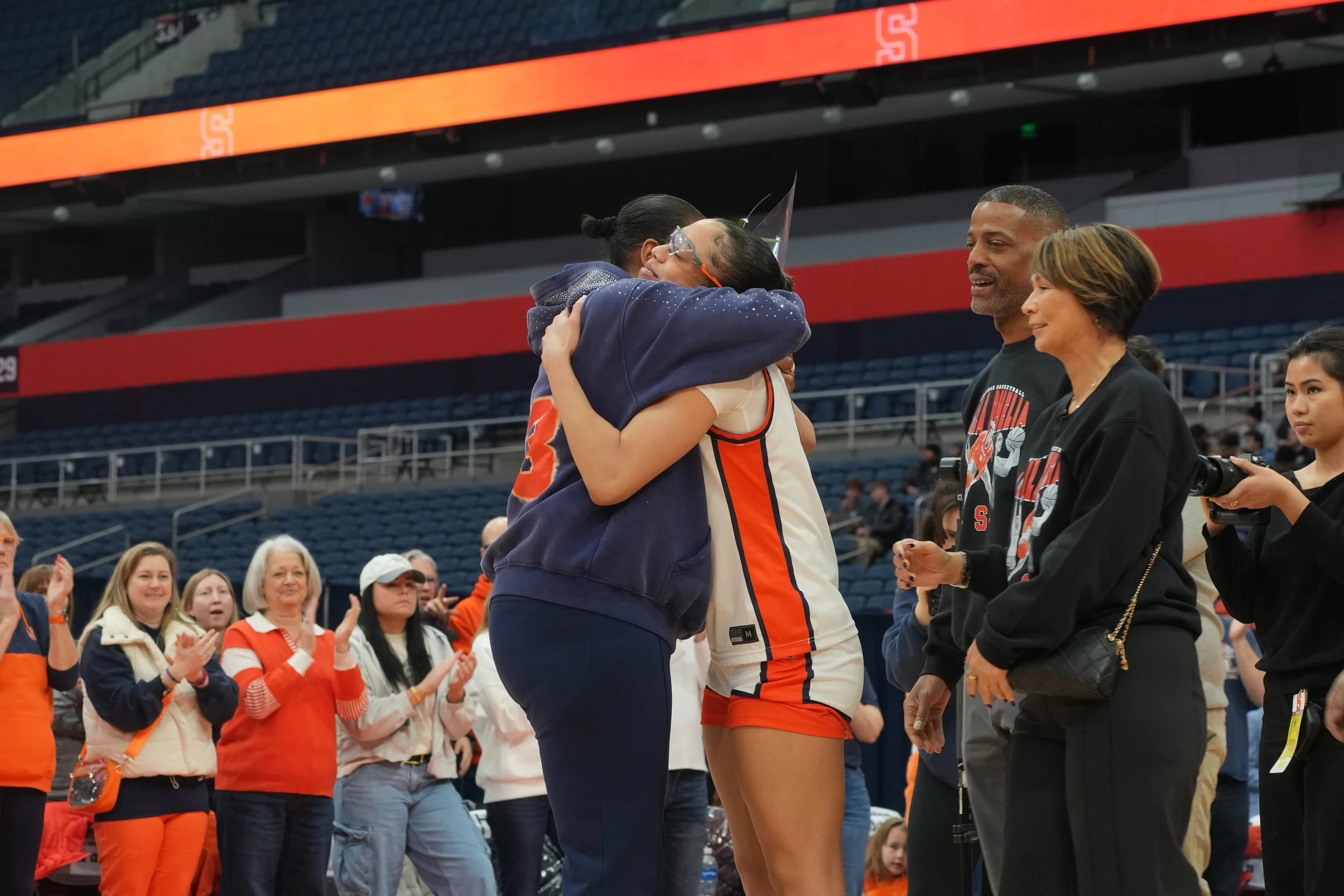 Two women hugging in a sports arena, surrounded by people clapping and smiling, with a basketball player in an orange and white uniform.