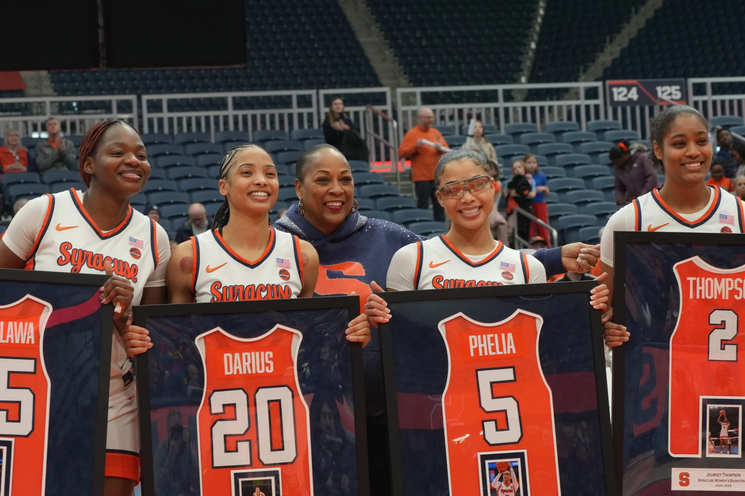 A group of five female basketball players and a woman in a navy blue hoodie are standing on a sports court, holding framed jerseys with their names and numbers. The players are wearing white Syracuse team jerseys, and the arena has mostly empty seats in the background.