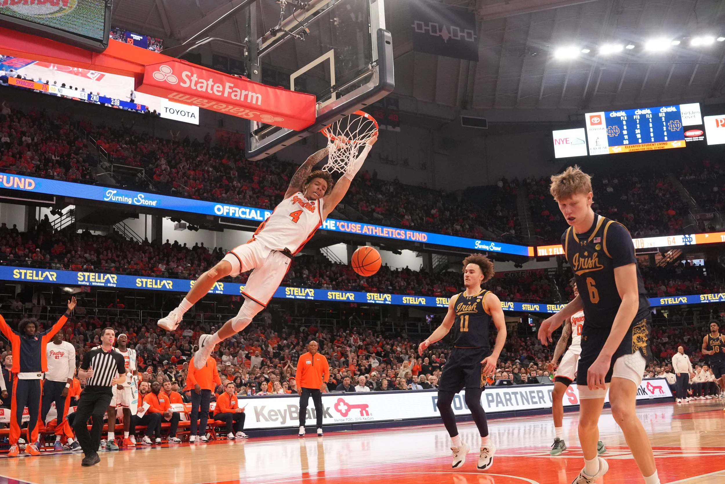 A basketball player in a white and orange uniform is dunking the ball into the basket during a game. Opposing players in dark blue and gold uniforms are on the court, with one looking down and another moving. The crowd is watching from the stands, and game advertisements are visible around the arena.