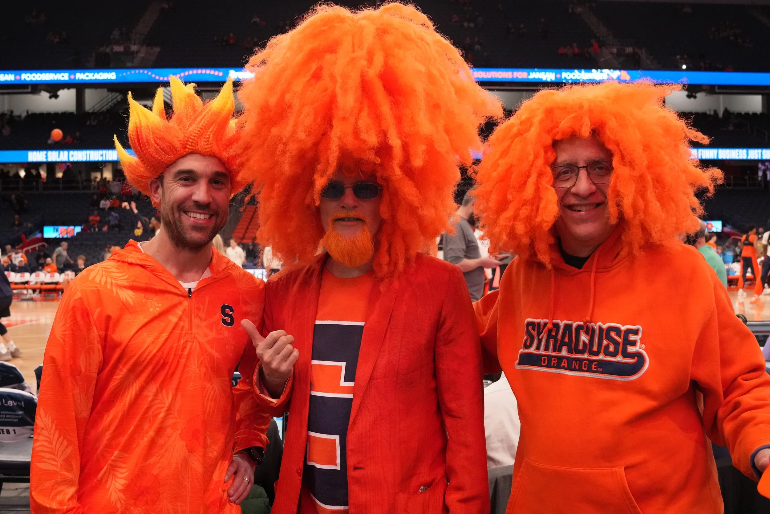 Three men dressed in orange clothing and wearing orange wigs at a sports arena. The man on the left is smiling and wearing an orange jacket with a letter 'S' on it. The man in the middle has a large orange wig, sunglasses, a beard, and is wearing a red blazer over a Syracuse University shirt. The man on the right is smiling, wearing glasses, an orange curly wig, and a Syracuse University hoodie.