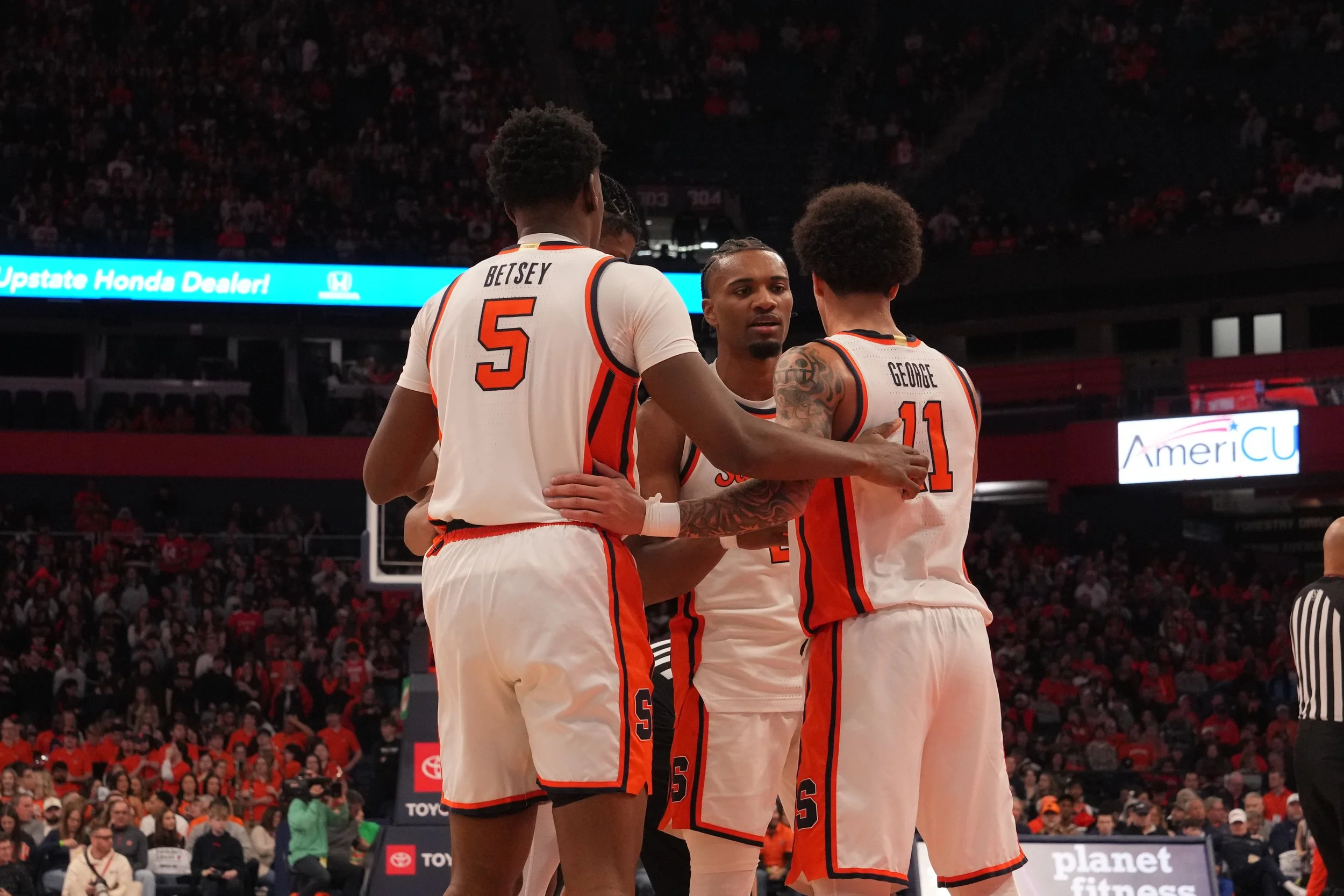 Four basketball players in white and orange uniforms standing together on the court during a game, with a crowded arena in the background.