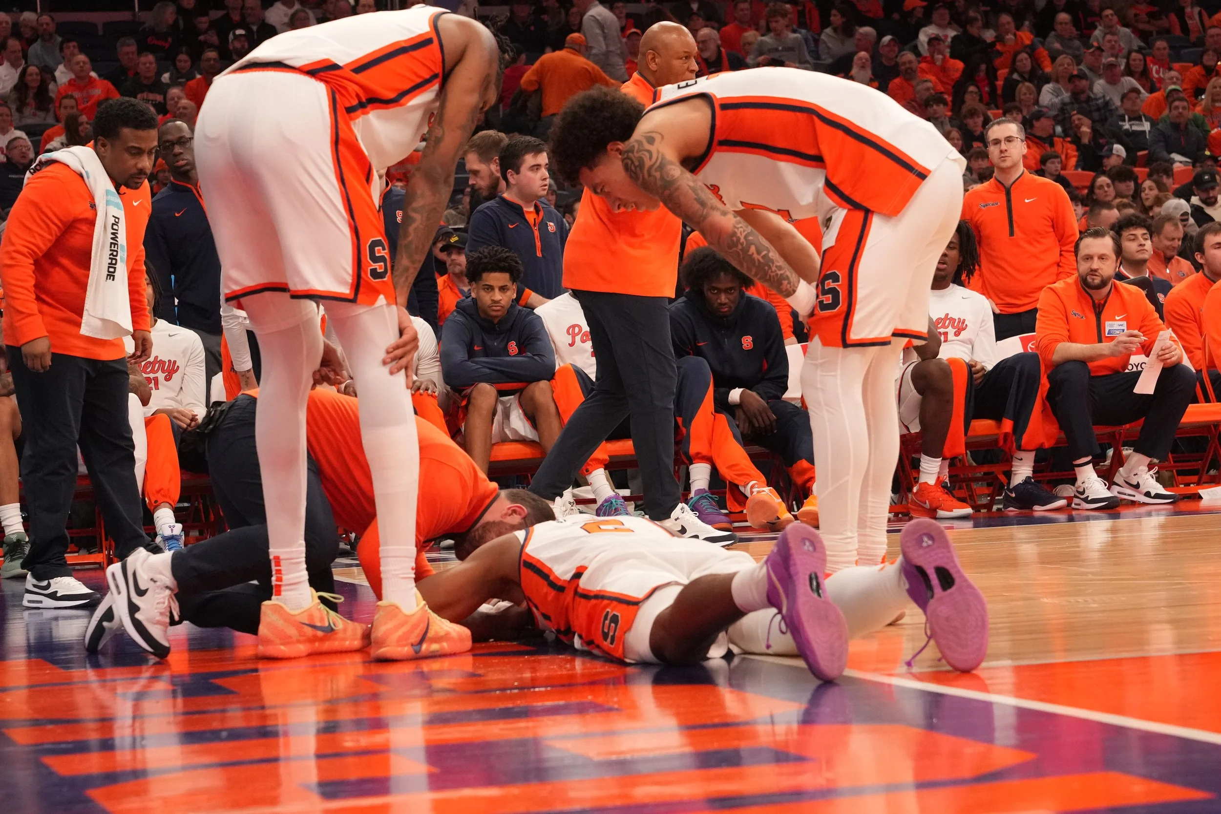 A basketball player is lying on the court, assisted by a staff member, while two teammates bend over him in concern during a game, with other players and spectators in the background.