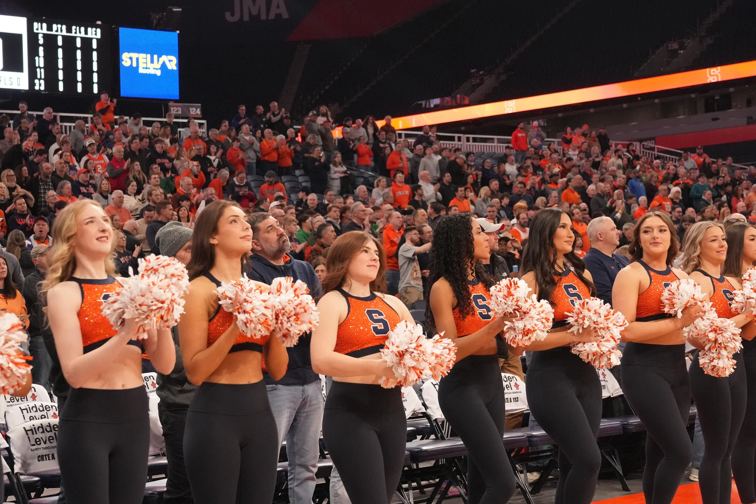 Cheerleaders holding pom-poms standing during a sports event at a stadium with a crowd in the background.