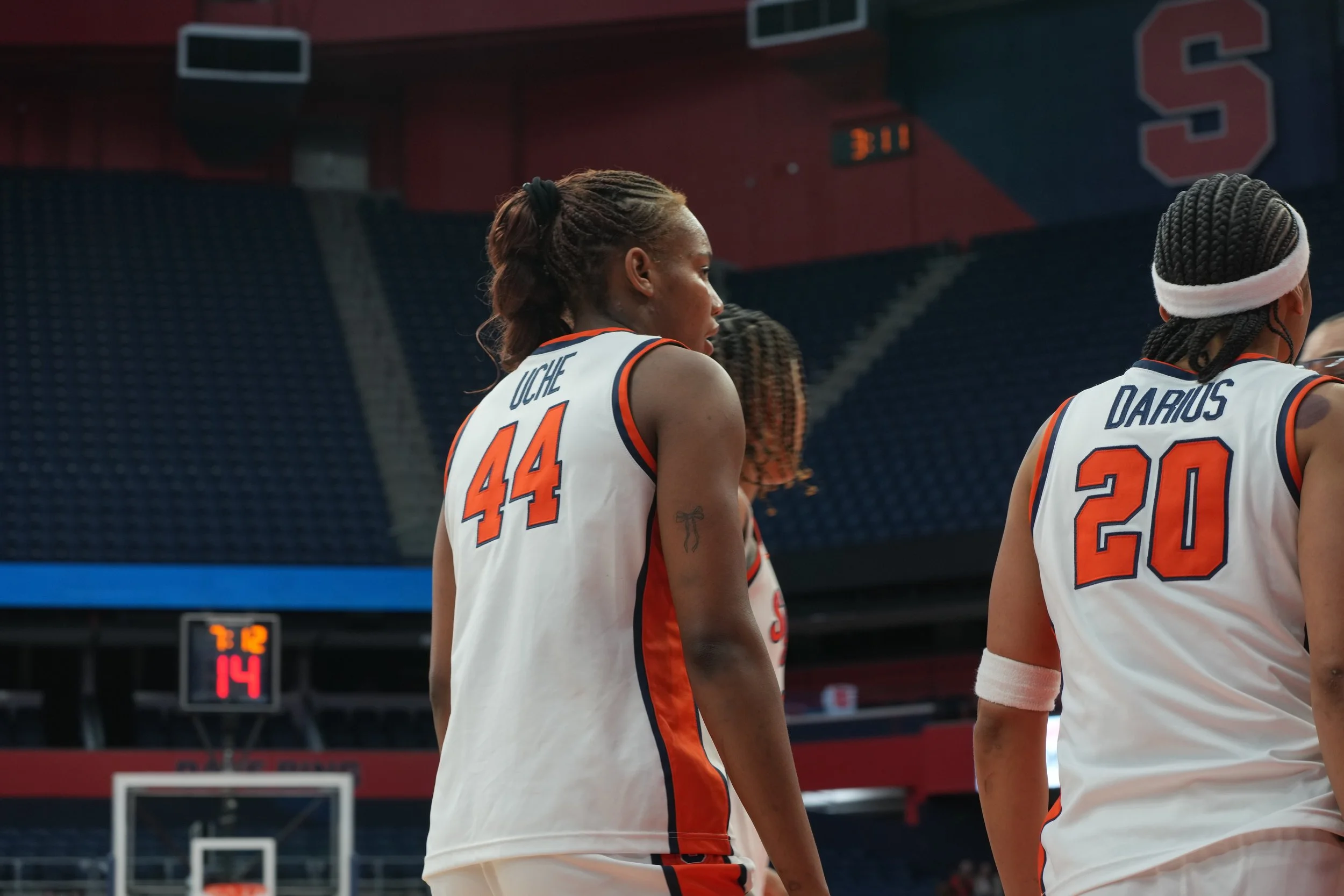 Two female basketball players in white jerseys with orange and blue accents on a basketball court, with a digital scoreboard and empty stands in the background.