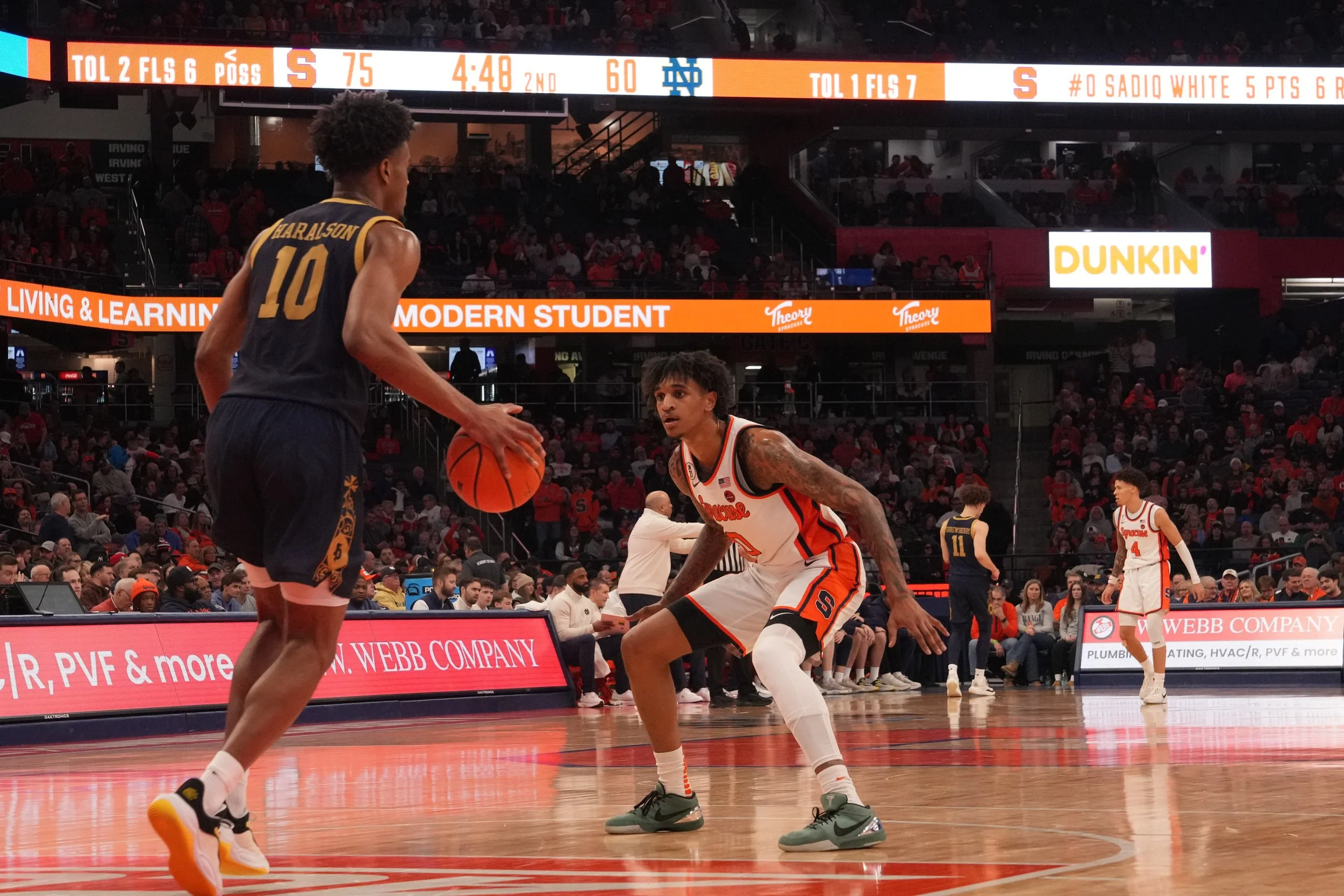 Two basketball players on the court during a game, one player in a dark jersey with the number 10 and the other in a white and orange jersey with the number 0, with a crowd watching from the stands and digital advertisements around the court.