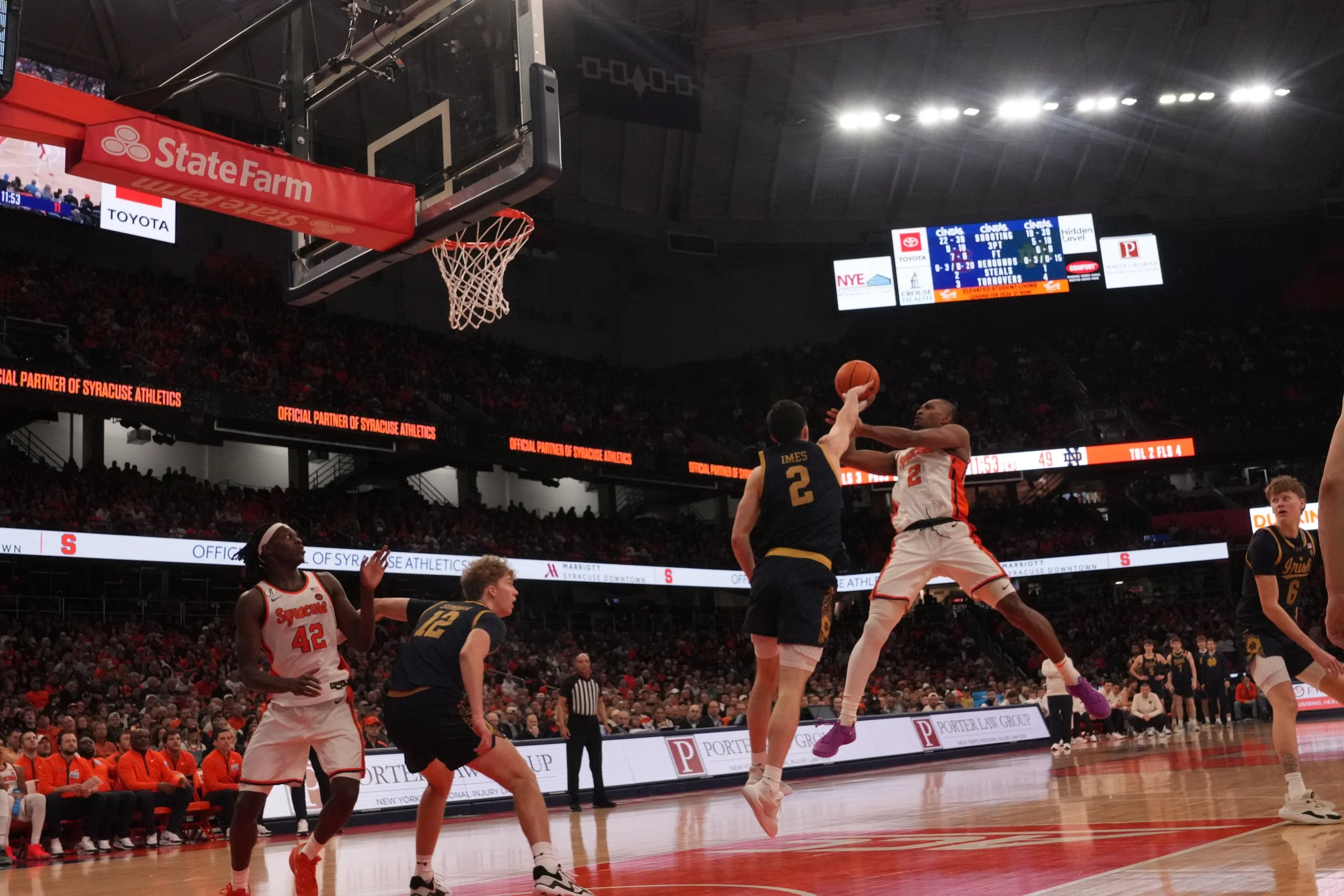 A basketball game in progress with players jumping and reaching for the ball near the hoop, with a crowd in the background, scoreboard, and advertisements visible.