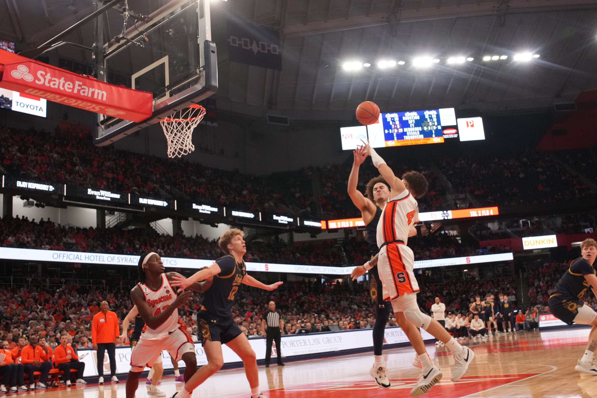 A basketball game in progress with players attempting a shot, one player in white jumping to score while a player in black defends. Others are nearby, and the crowd is visible in the background.