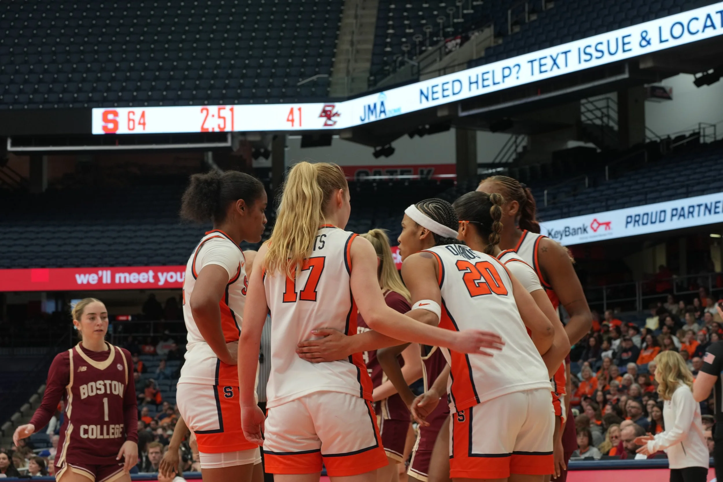 Women's basketball team huddling on the court during a game, with a player from Boston College standing nearby.