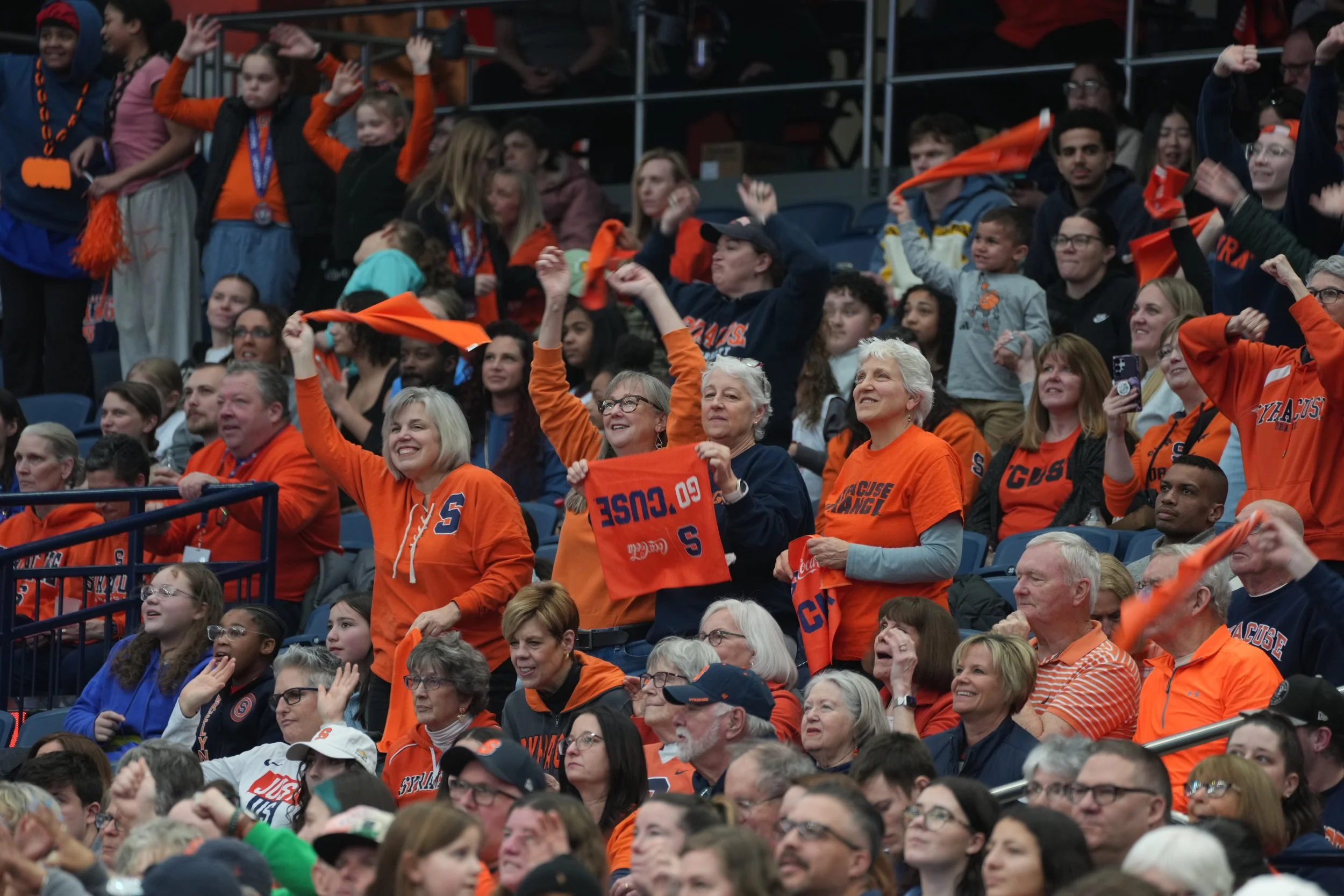 Crowd of sports fans at an indoor arena, many wearing orange and blue Syracuse University apparel, cheering and waving towels.