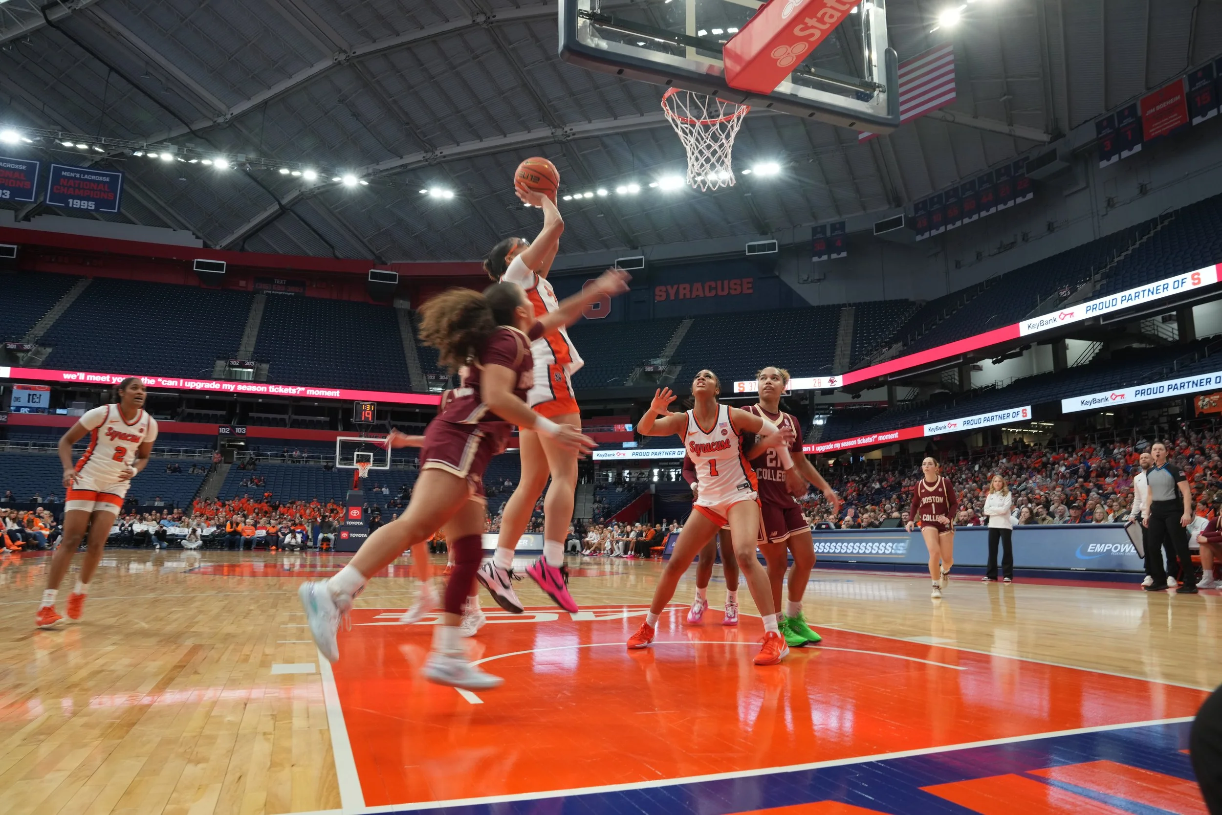 Women playing basketball on an indoor court. One player in a white and orange jersey is jumping to shoot or block the ball near the hoop, while another in a maroon jersey attempts to defend. Other players and spectators are visible.