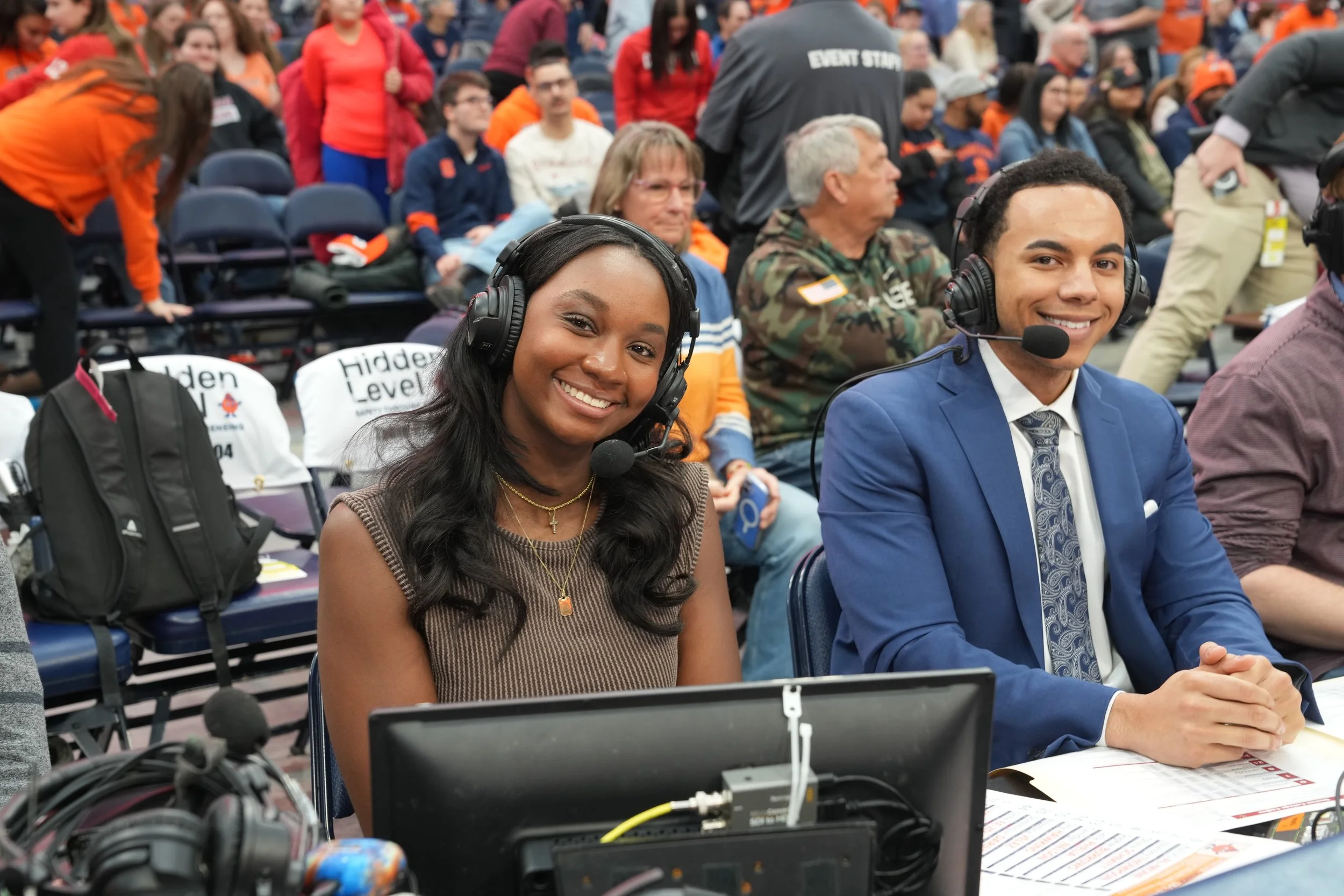 Two broadcast announcers, sitting at a table with headphones and microphones, smiling at the camera, surrounded by a crowd of spectators at an indoor event.