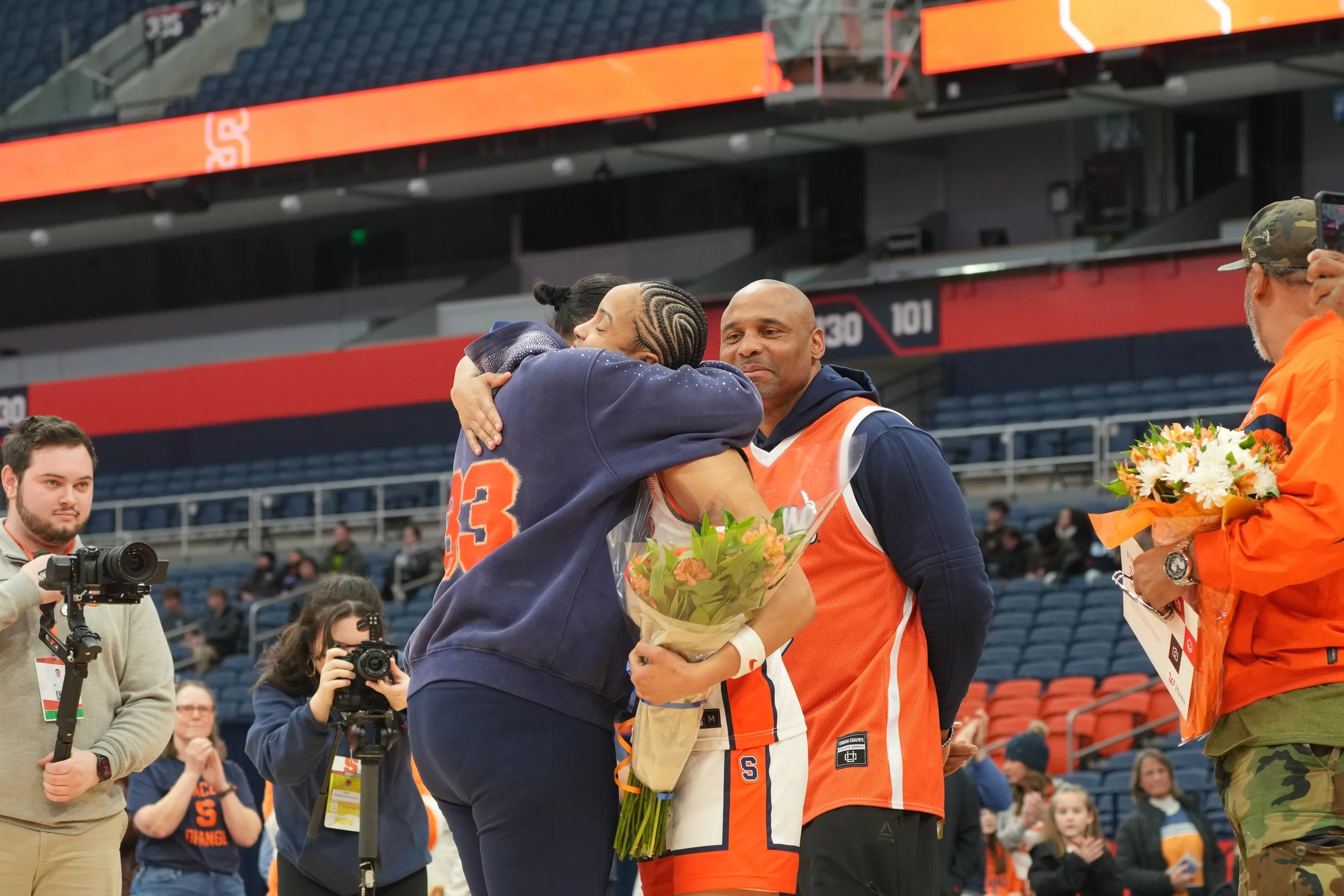 Two basketball players hugging, one handing a bouquet of flowers to the other, surrounded by photographers and spectators in a stadium.