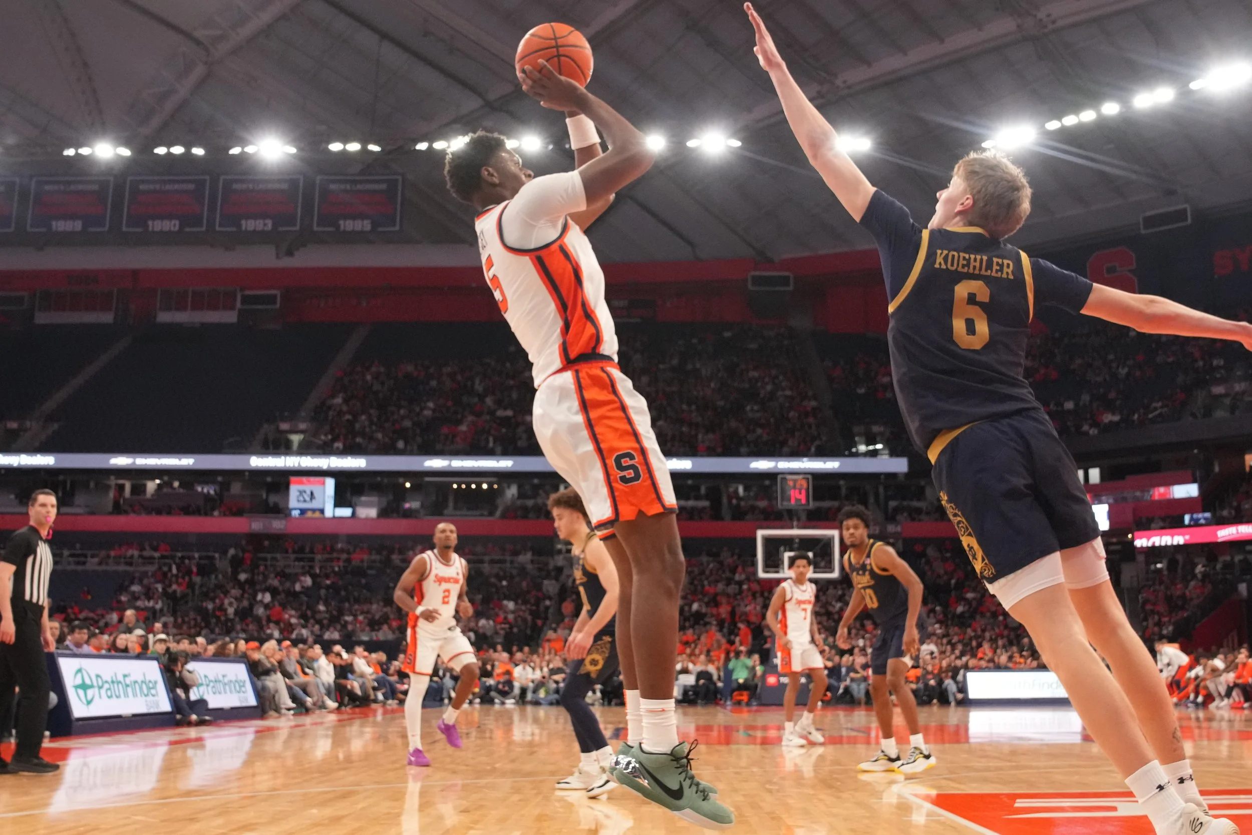 A basketball game with a player in a white and orange uniform jumping to shoot the ball while an opposing player in a navy blue and yellow uniform attempts to block the shot. The game is indoors with a crowd in the background.