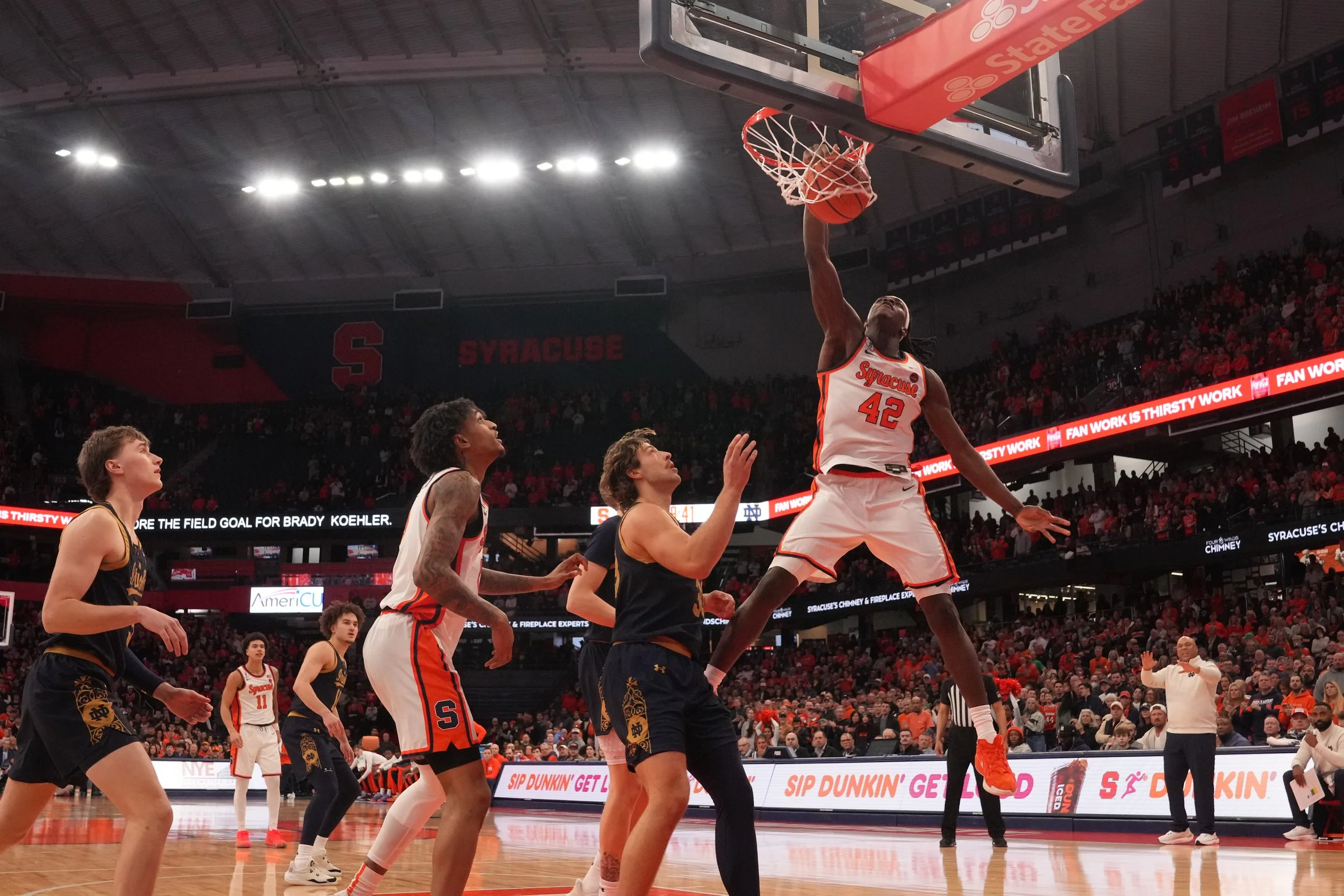 A basketball player in a Syracuse uniform, number 42, is dunking the ball during a game with players from both teams watching and cheering on the court at Syracuse's arena.
